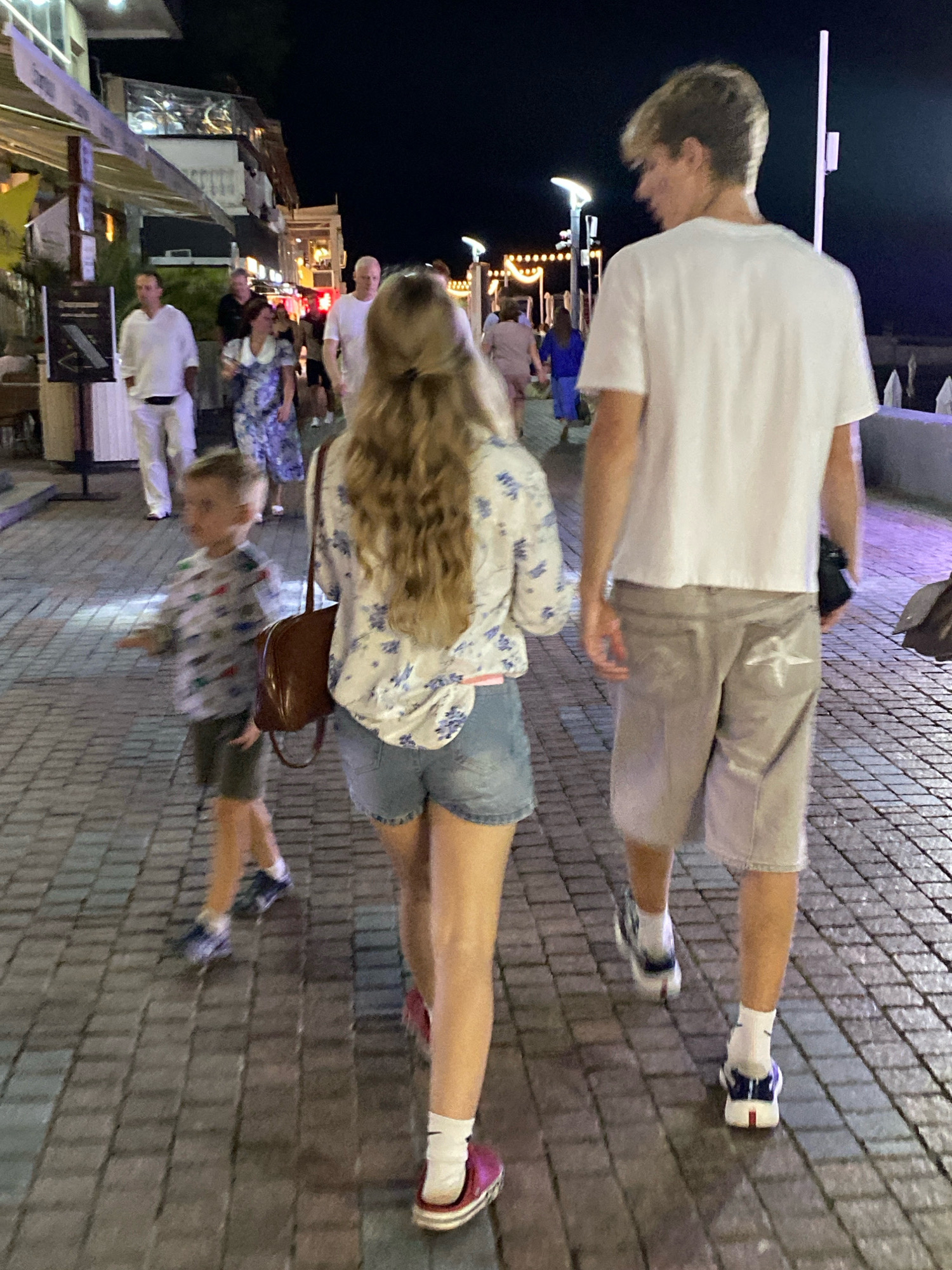 In Sochi, Russia, at night, four people are walking on a cobblestone street. The foremost person is a young boy wearing a long-sleeved shirt with a pattern of colorful cars and grey shorts. He is being followed by a young woman with long, blonde, wavy hair, wearing a floral-patterned blouse and denim shorts. She carries a brown handbag over her shoulder. Behind her, a young man in a white t-shirt and light-colored shorts walks away from the camera. In the background, a man in a white t-shirt and white pants is visible, as are other pedestrians and illuminated storefronts along the street. The scene depicts a pedestrian promenade at night.