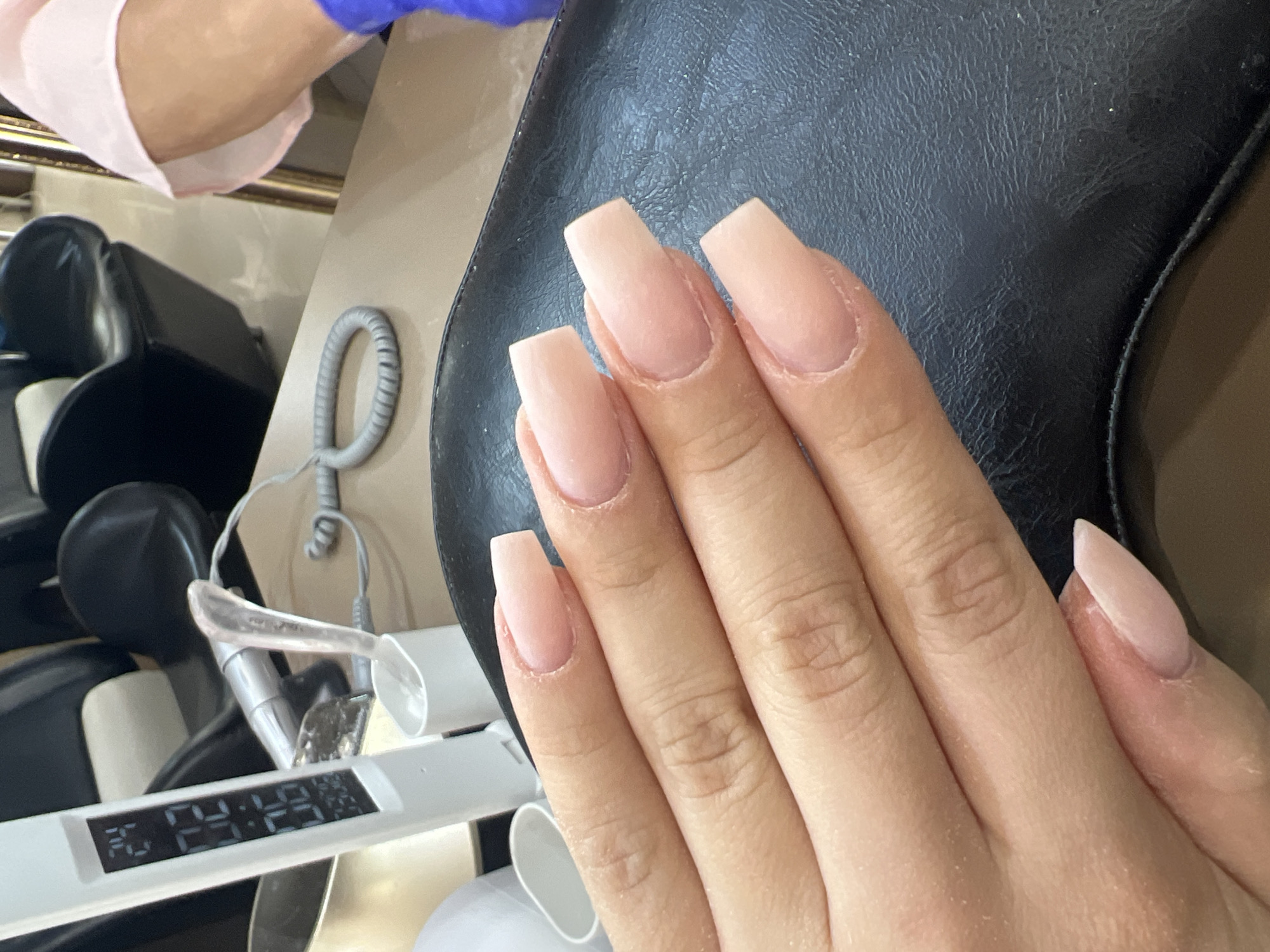 This close-up image shows a person's right hand with artificial nails, likely taken in a nail salon in Plovdiv, Bulgaria. The hand is positioned against a dark, textured surface, which appears to be part of a salon chair. The nails are a natural, light nude pink color with a matte finish, suggesting they are either freshly applied or in the process of being completed. Most nails feature a coffin or ballerina shape, while the pinky finger's nail has a more almond or stiletto tip. Fine white dust, possibly from filing or buffing, is visible on the fingers and around the cuticles.
In the upper left of the frame, a segment of a nail technician's arm can be seen, wearing a blue glove and a pink sleeve, indicating an ongoing nail service. To the lower left, a white rectangular device with a digital display shows the numbers "30:29:29". This is likely a timer or a temperature and time readout associated with nail curing or drying equipment. A coiled gray cable is also visible on a light-colored surface in the mid-ground. The indoor setting, specialized equipment, and the state of the nails clearly point to an active beauty treatment session. The precise time of day is not discernible from the lighting.