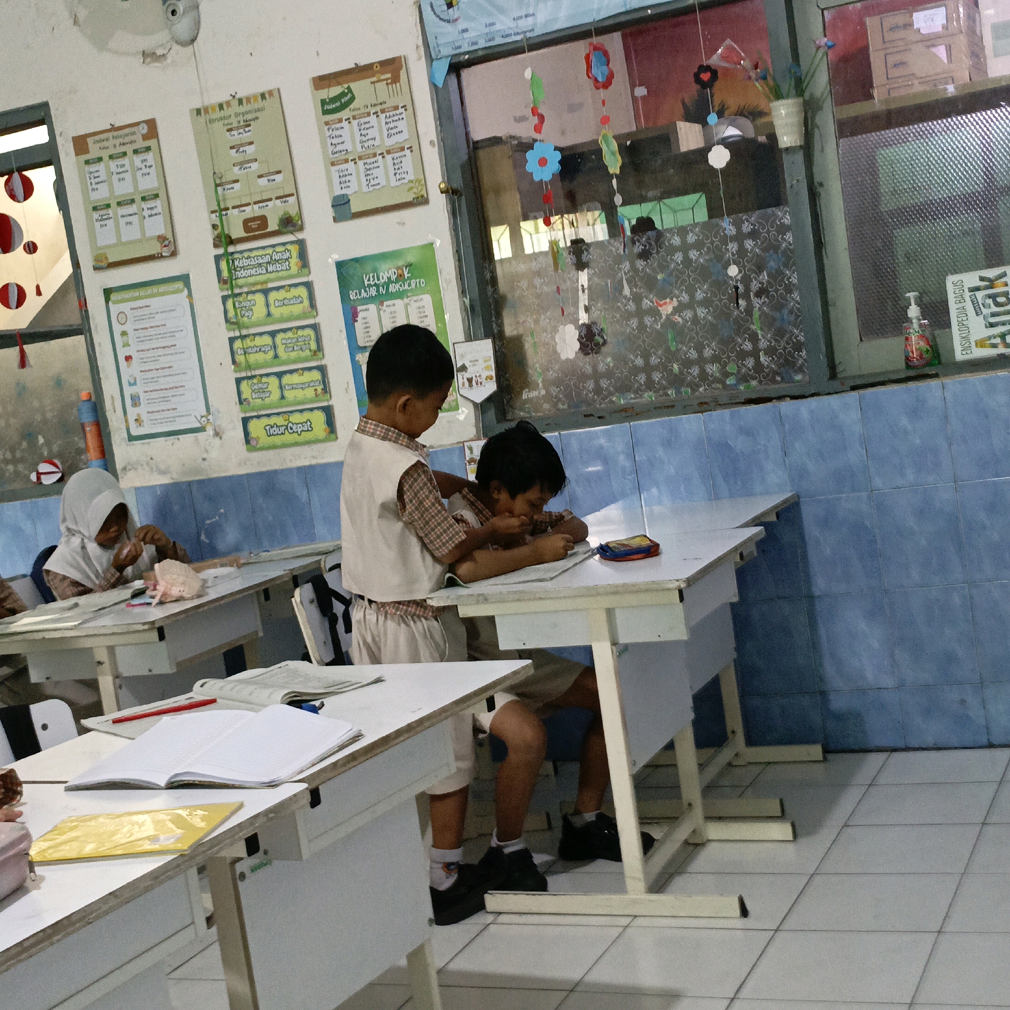 The image depicts a classroom setting, likely in an elementary school, with two young boys in uniform seated at desks. One boy, wearing a light-colored vest over a checkered shirt and white pants, stands behind another boy who is hunched over his desk, appearing to be writing or reading. The classroom walls are adorned with colorful educational posters. One poster displays a "Struktur Organisasi Kelas IV A" (Organizational Structure of Class IV A), while another details "Kebiasaan Anak Indonesia Hebat" (Habits of Great Indonesian Children). A third poster, "Jadwal Pelajaran" (Lesson Schedule), is also visible.
A girl in a white hijab is seated at a desk in the foreground, to the left of the frame, also engaged in an activity at her desk. Desks are white with wooden frames and legs. The floor is tiled in a light color. The environment appears to be a typical Indonesian classroom, potentially located in Salatiga, Indonesia, given the context provided. Natural light streams in from a window on the left side of the image, and a fluorescent light fixture is visible on the ceiling. Decorative elements, such as paper cutouts shaped like flowers and circles, hang from the ceiling and window frames. A hand sanitizer bottle is visible on a ledge near the window.