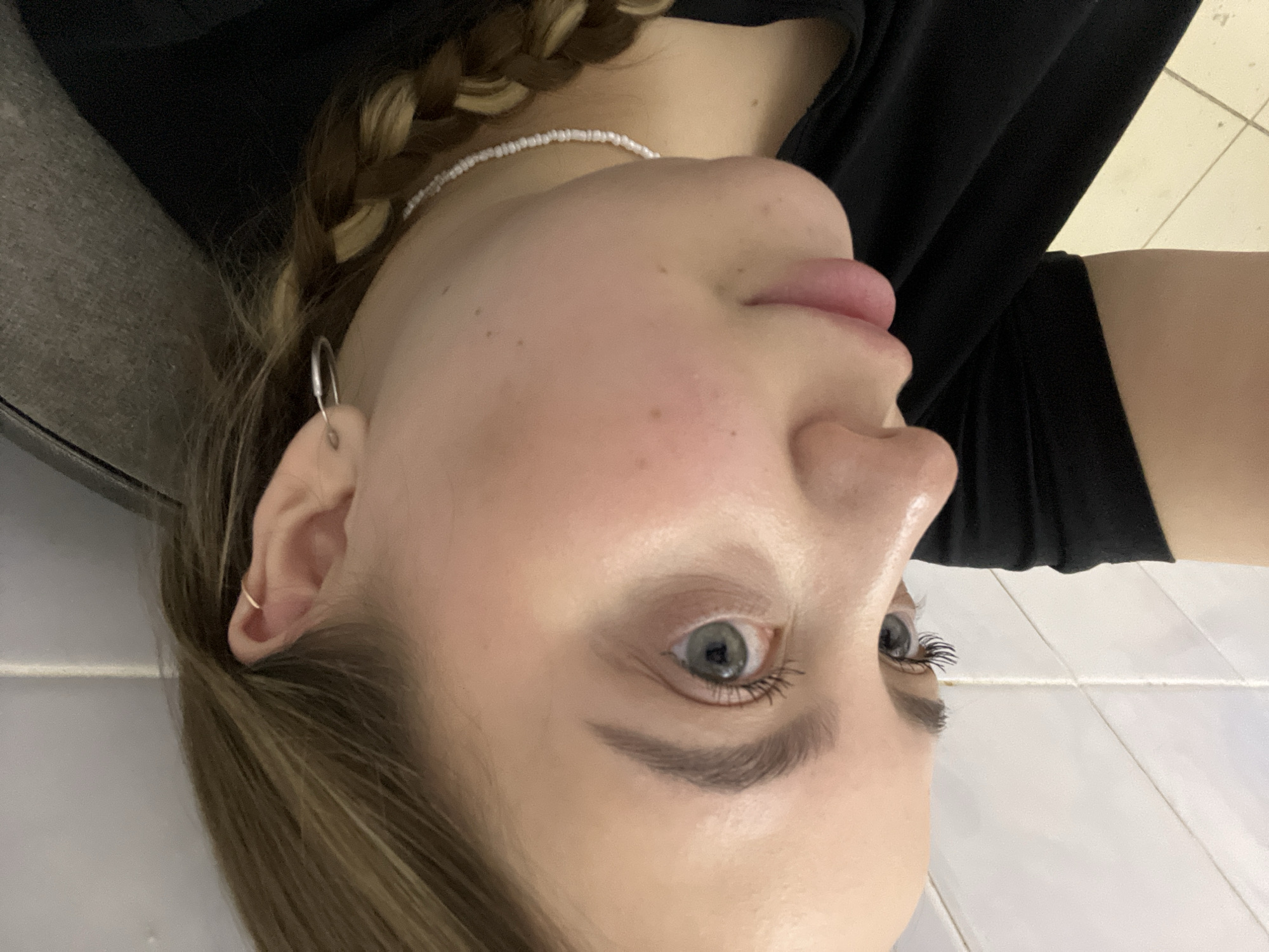 A close-up, tilted portrait of a young woman with light skin and blue eyes. She is wearing a black top. Her hair is braided and she has a small silver earring in her left ear. A beaded white necklace adorns her neck. The background appears to be a tiled surface. The location of the photo is not identifiable as Novopokrovskaya, Russia based on the image.