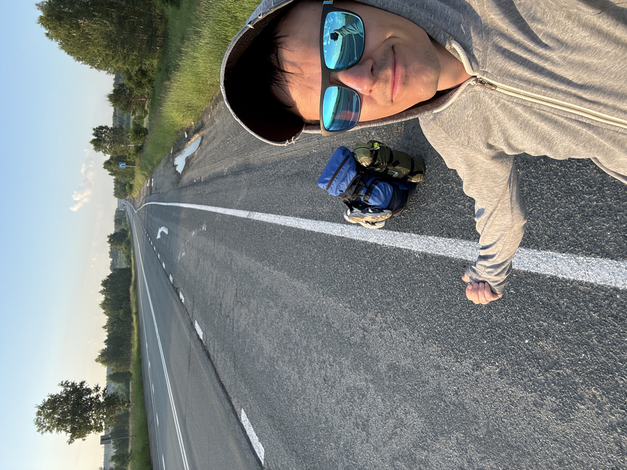 A young man with dark hair, wearing a grey hooded sweatshirt and sunglasses, stands on the side of a paved road in a rural setting. He is holding his right arm out to the side with his fist clenched, in what appears to be a hitchhiking gesture. Next to him on the road is a blue and green backpack. The road has white lines marking the lanes and a shoulder. The environment consists of green grass and trees on either side of the road, with a clear blue sky overhead. The scene suggests a journey or travel. The location is identified as Unknown, Russia.