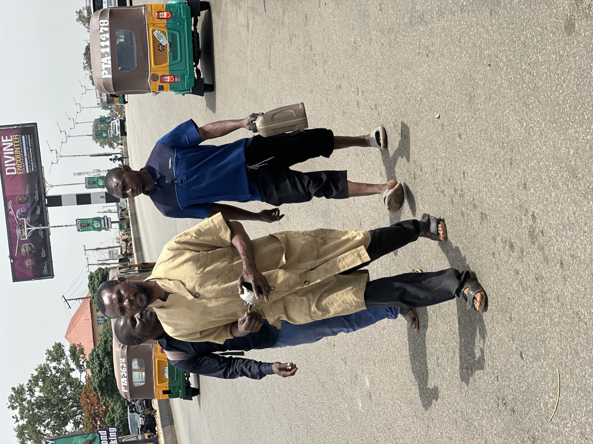 The image shows a street scene in Jos, Nigeria, during daylight. Two men are standing on a paved road. One man is wearing a khaki overcoat and black pants. The other man is wearing a blue shirt and black shorts and holding a light-colored container. Both men are wearing sandals. In the background are auto rickshaws and a billboard advertising a "Divine Encounter" event. Streetlights and other vehicles are visible in the background.