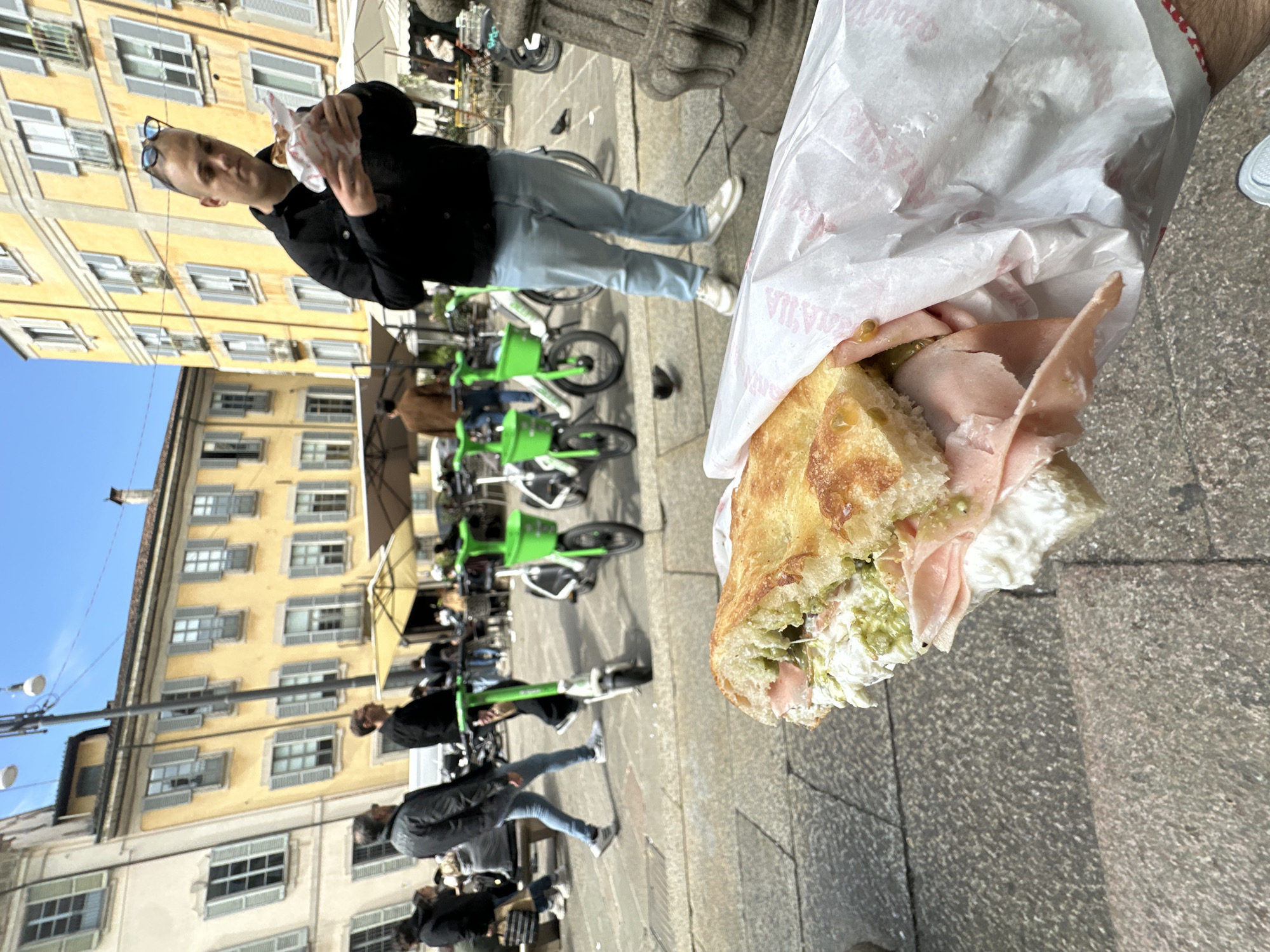 An outdoor, eye-level shot captures a man holding a large sandwich in the foreground, set against an urban backdrop in what appears to be Milan, Italy. The image is rotated, showing the man standing on a paved street.
The man, positioned towards the left side of the frame, wears a black jacket and light blue jeans, with sunglasses resting on his head. He holds a sandwich, likely a focaccia, wrapped in white paper. The sandwich is generously filled with slices of pink cured meat (resembling mortadella), a green spread (possibly pesto or a similar condiment), and some white creamy ingredient.
In the background, tall, multi-story buildings with yellow and light beige facades line the street. These buildings feature numerous windows with light-colored shutters, characteristic of European architecture. Below the buildings, several bright green share-bikes (Lime bikes) are parked along the sidewalk, adding to the urban scene. Other pedestrians are visible walking further down the street, blurred in the background.
The setting suggests a sunny day with a clear blue sky, indicating good weather. The overall activity centers around a casual moment of enjoying street food in a bustling city environment. Part of a red border and some indistinct white text (possibly "NATALIA" or "MATILDA") are visible on the paper wrapping the sandwich. The paved ground is made of grey stones or concrete.