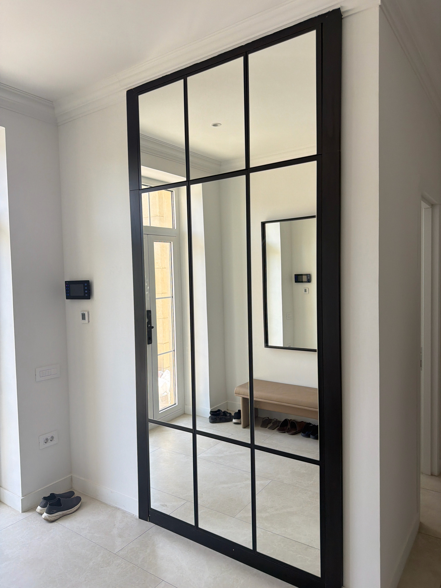 A tall, black-framed mirror with multiple panels occupies the center of the frame, reflecting a hallway with a door and a bench. The mirror is installed on a white wall that features crown molding at the ceiling. To the left of the mirror, near the bottom, a pair of dark sneakers rests on a light-colored tiled floor. Further up the wall, a black electronic device with buttons and a small white thermostat are mounted. A light switch panel is visible lower down on the wall. Reflected in the mirror, a light-colored bench is present, with a row of shoes placed in front of it, including sandals and dress shoes. A rectangular mirror is also visible in the reflection, mounted on the wall behind the bench, showing another electronic device. The floor is tiled with large, light-colored square tiles. The overall scene suggests an interior hallway or entryway, possibly in a residential or commercial building. The mention of Sabunchu Raion, Azerbaijan, is external information and not derivable from the image itself.
