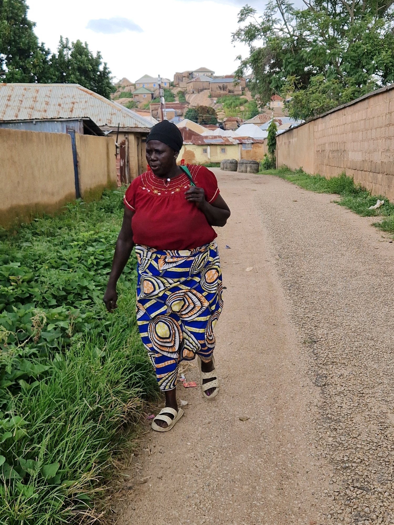An outdoor, full-shot, medium-angle, eye-level, medium-distance, vertical, full-color, daylight, natural-light photograph shows a Black woman walking on a gravel road. The woman is wearing a red t-shirt, a blue, yellow, and white patterned skirt, a black headscarf, and white slide sandals. She is carrying a green bag over her left shoulder and holding a green object in her right hand. The woman is facing right, looking forward, with her mouth open. She is walking past a patch of green grass and foliage on the left and a brown brick wall on the right. In the background, there are several buildings with corrugated metal roofs and houses on a hill. The sky is cloudy.