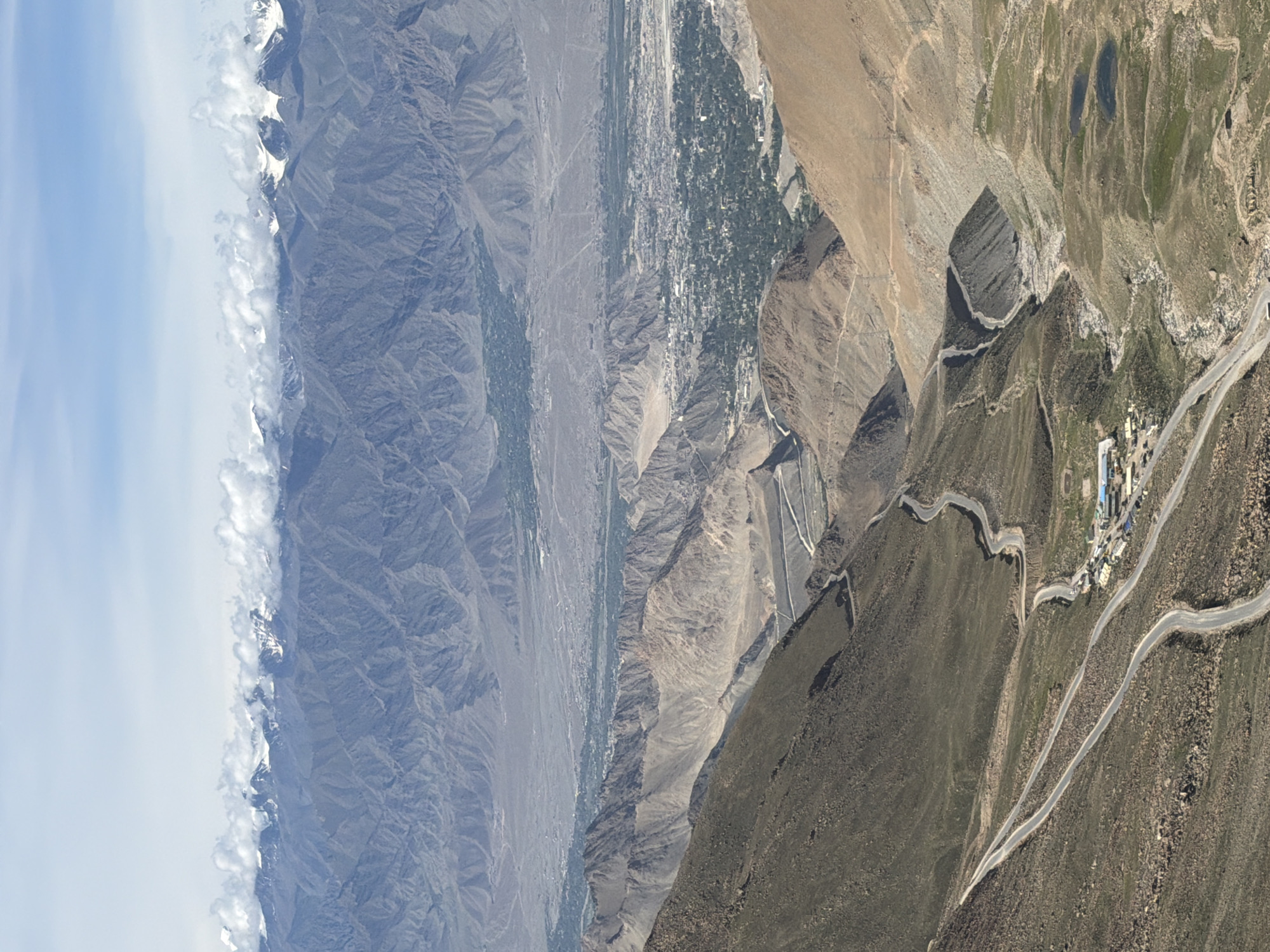 The image captures a high-altitude, panoramic view of the city of Leh, India, nestled within a rugged, mountainous landscape. In the foreground, a winding road snakes its way down a barren, rocky slope, disappearing into the valley below. The road appears to be a major thoroughfare, with at least two lanes visible in some sections, indicating significant travel infrastructure.
Further down the mountain, the road leads to a small cluster of buildings, likely a settlement or a rest stop, marked by several structures and parked vehicles. This area appears to be a point of transition between the harsh, arid mountains and the more fertile valley.
Beyond this, the vast expanse of Leh city unfolds. The city is characterized by a dense collection of buildings, appearing as a tapestry of gray and white structures against the green patches of vegetation that signify irrigated areas. The city is spread across the valley floor, surrounded by more undulating, arid mountains that recede into the distance.
The background is dominated by a majestic range of snow-capped mountains, their peaks piercing the sky. Wisps of clouds cling to these formidable peaks, adding a dramatic flair to the already breathtaking scenery. The sky above is a clear, bright blue, punctuated by scattered white clouds, indicative of a fair-weather day. The overall scene depicts a harsh yet beautiful natural environment, with human settlement strategically placed within the valley, a testament to the resilience of life in such demanding conditions.