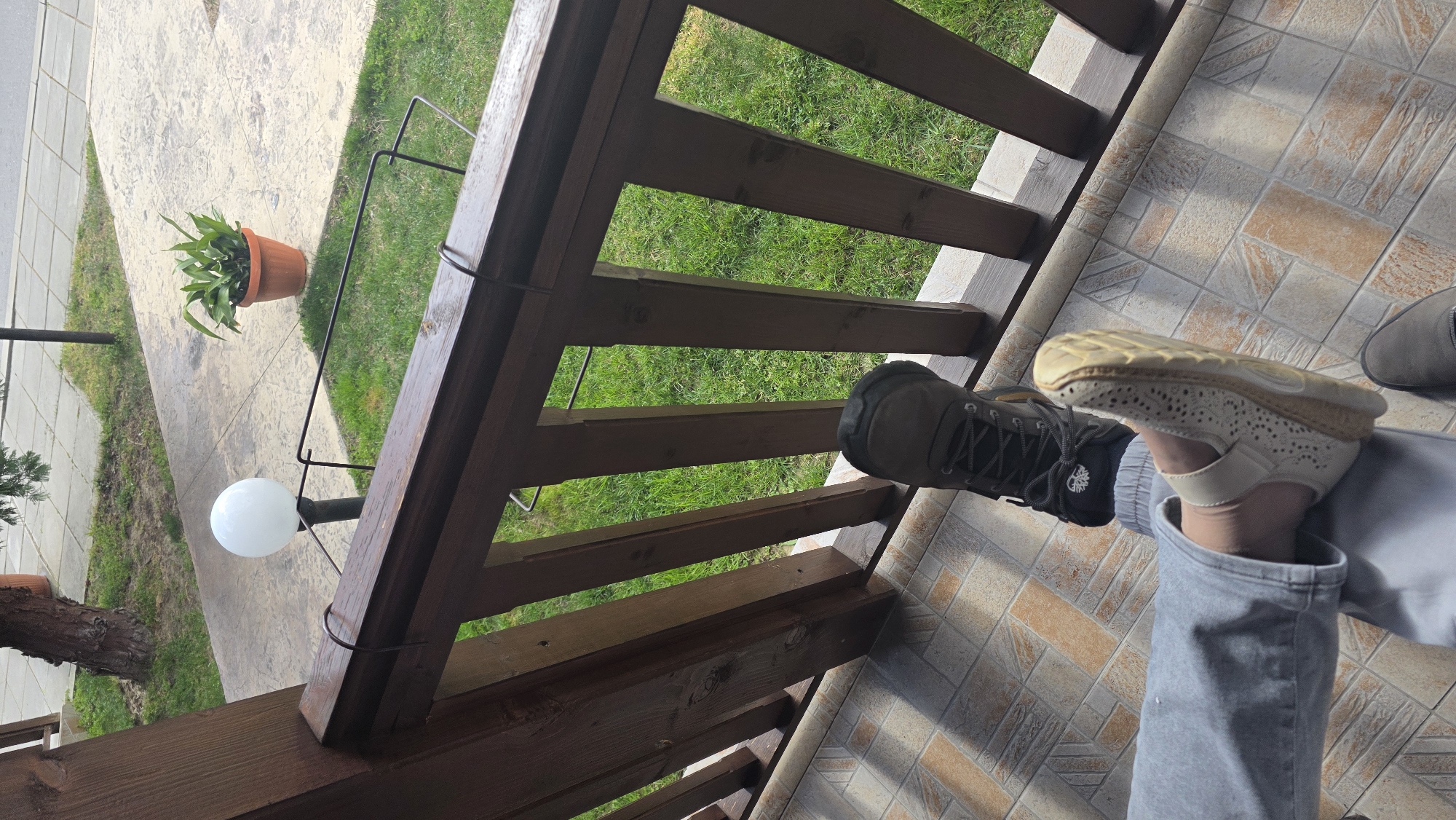 This image depicts an outdoor patio or balcony located in the city of Rila, Bulgaria. The foreground shows a tiled floor composed of light brown and beige square tiles. A dark brown wooden railing, featuring a horizontal top rail and vertical balusters, crosses the mid-ground, with metal hangers attached to the top rail. Two sets of feet are visible on the tiled surface: one foot wears a dark grey hiking boot, and another, clad in light grey sweatpants, wears a light beige perforated sandal. A third dark grey slip-on shoe is partially visible at the bottom. Beyond the railing, a strip of vibrant green grass is visible, bordered by a light grey patterned concrete pathway. On the pathway, a spherical white outdoor lamp on a dark pole stands, accompanied by a terracotta pot containing a green leafy plant. A tree trunk is partially visible on the far left. The scene is illuminated by natural light, casting shadows on the tiled floor.