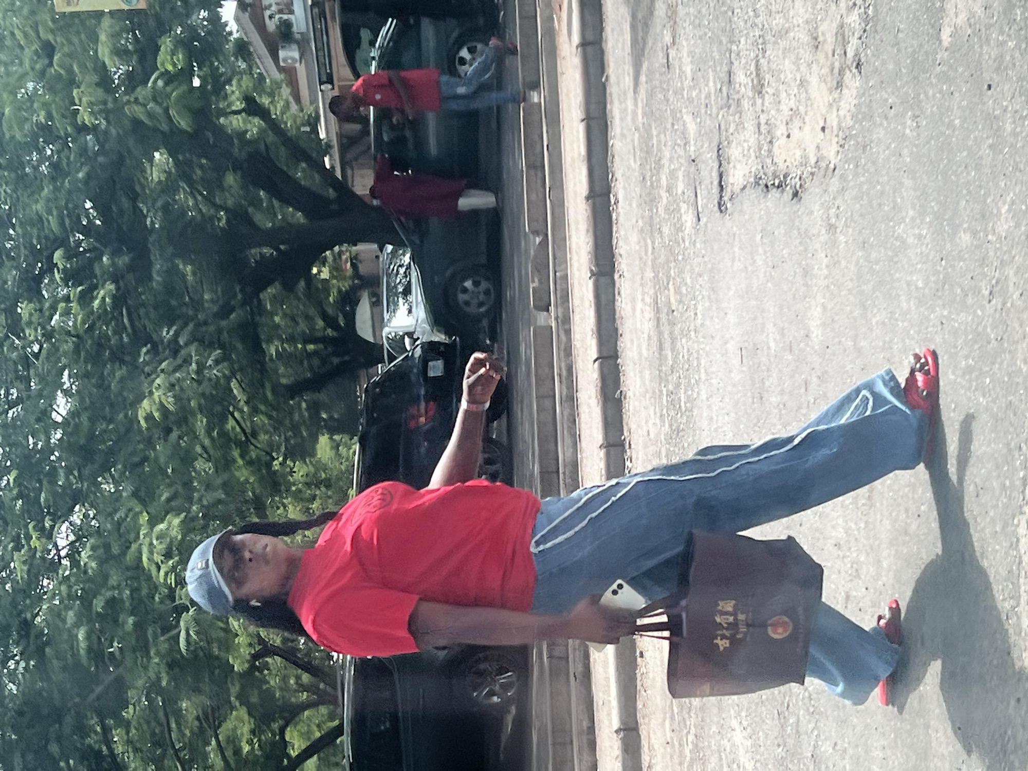 A woman stands on a paved road, potentially a sidewalk, in what appears to be an urban environment. She is wearing a red t-shirt, blue jeans with white stripes, a blue cap, and red shoes. She carries a cell phone in one hand and a brown tote bag with what looks like Asian characters on it in the other. In the background, there are parked cars and trees. There are pedestrians in the background and it is the city of Abuja, Nigeria.