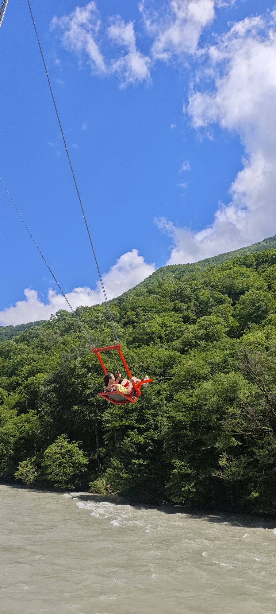 This vertical shot captures a thrilling outdoor activity in a scenic mountainous landscape. The central subject is a person, likely a woman, seated in a vibrant red swing suspended high above a river. She is wearing a yellow top and light-colored bottoms, and is holding a mobile phone in her hands, possibly recording or taking a picture. Her posture appears relaxed as she hangs suspended by multiple thick metal cables.
The setting is characterized by a dramatic natural environment. In the background, a steep, densely forested mountain rises under a bright blue sky adorned with scattered white clouds. The trees on the mountainside are lush and green, indicating a warm, possibly summer, season. Below the swing, a wide river flows, its water appearing a turbid light brown or greyish-white, suggesting a strong current or glacial melt. The riverbank on the far side is also heavily forested.
The image conveys a sense of adventure and exhilaration, with the person enjoying the panoramic views from a considerable height. The bright daylight and clear sky suggest excellent weather conditions for such an activity. The scene is set in a picturesque natural area, specifically Bzypta, Abkhazia. No visible text is present in the image.