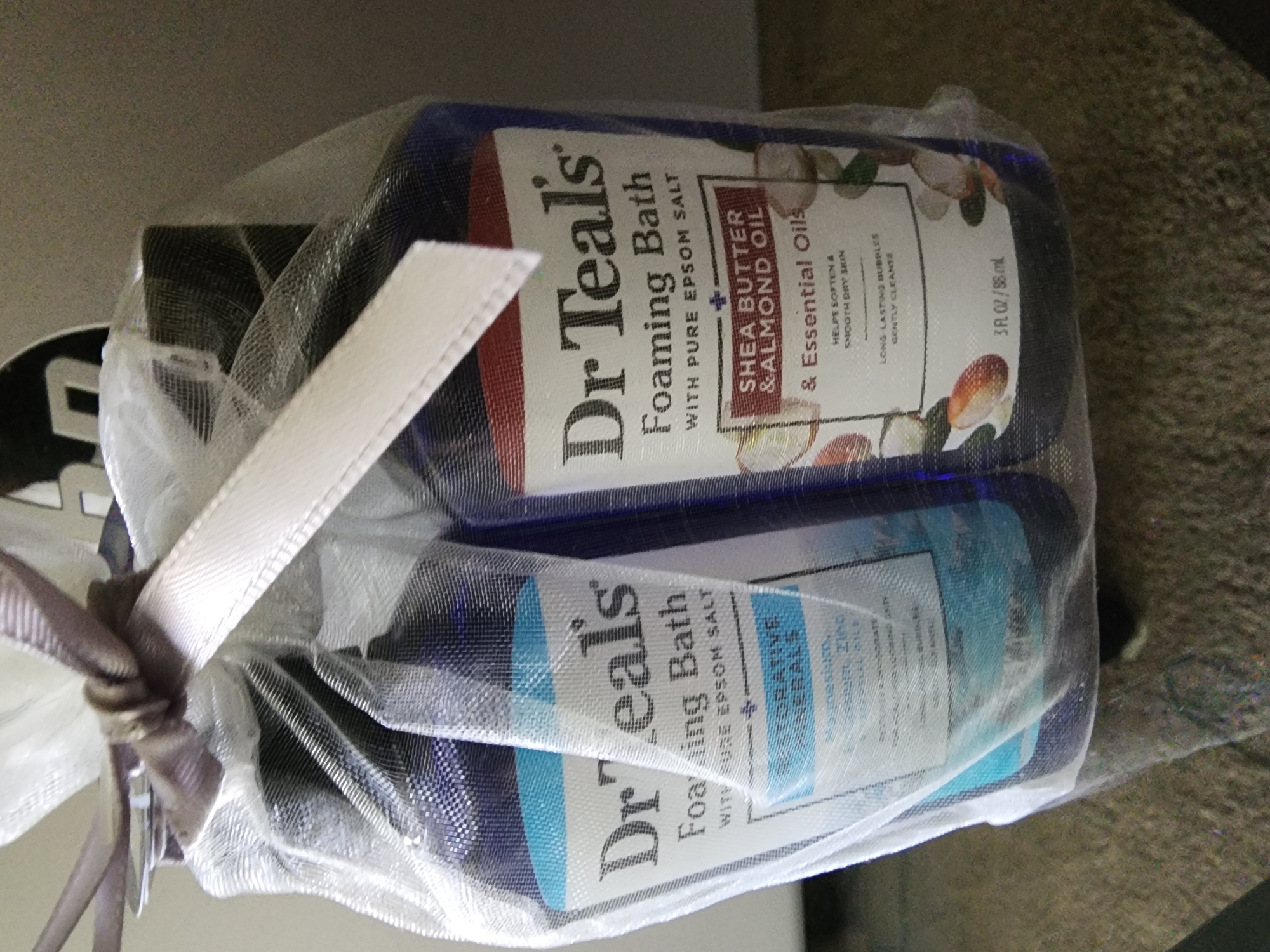 This is a close-up, eye-level shot of two Dr. Teal's Foaming Bath products tied together with a sheer white bag and a brown and white ribbon. The bottles are dark blue and have white labels with black text. The product on the left appears to be "Dr Teal's Foaming Bath with Pure Epsom Salt" and "Restorative Minerals". The product on the right is "Dr Teal's Foaming Bath with Pure Epsom Salt Shea Butter & Almond Oil & Essential Oils". Both bottles have a volume of 3 FL. OZ./88 mL. The background is blurred and appears to be an indoor setting, possibly a home. The location is Lower Paxton Township, United States of America.