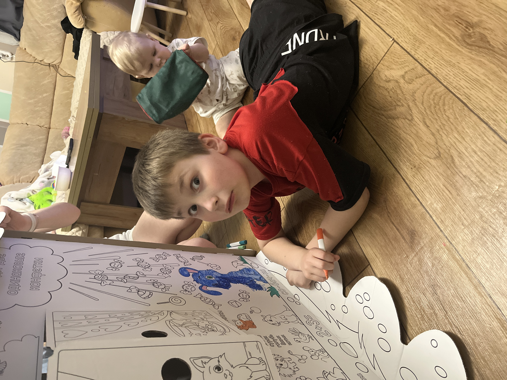 Scene: An indoor shot shows two children, a drawing book, and other objects.
People: One child, a boy, is lying on the floor facing the camera and is coloring. Another child is in the background.
Subjects: The primary subject is the boy coloring. Other subjects include the second child, the drawing book, and the markers.
Objects: The boy is holding an orange marker and coloring in a large cardboard drawing book filled with images to color. In the background, a younger child is holding a green bag. Other objects in the scene include markers, a couch, and a wooden floor.
Actions: The boy is actively coloring in the drawing book. The younger child is holding a bag.
Interactions: The boy is interacting with the drawing book and the markers. The second child seems to be playing with a bag.
Environment: The scene appears to be inside a home or a living room, with a wooden floor, a couch, and other furniture. The location is not specified as Oryol-Izumrud, Russia, based on the visible elements in the image.
