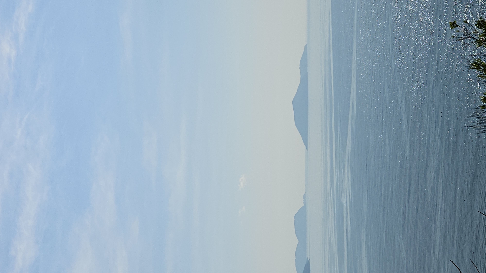 The image presents a hazy view of the sea and sky near Nam Natural Heritage Area Trang, Vietnam. Silhouetted landmasses are visible in the distance, suggesting islands or peninsulas. The sea surface reflects light with a shimmering effect. Vegetation is present on the right side of the image. The sky is lightly clouded.