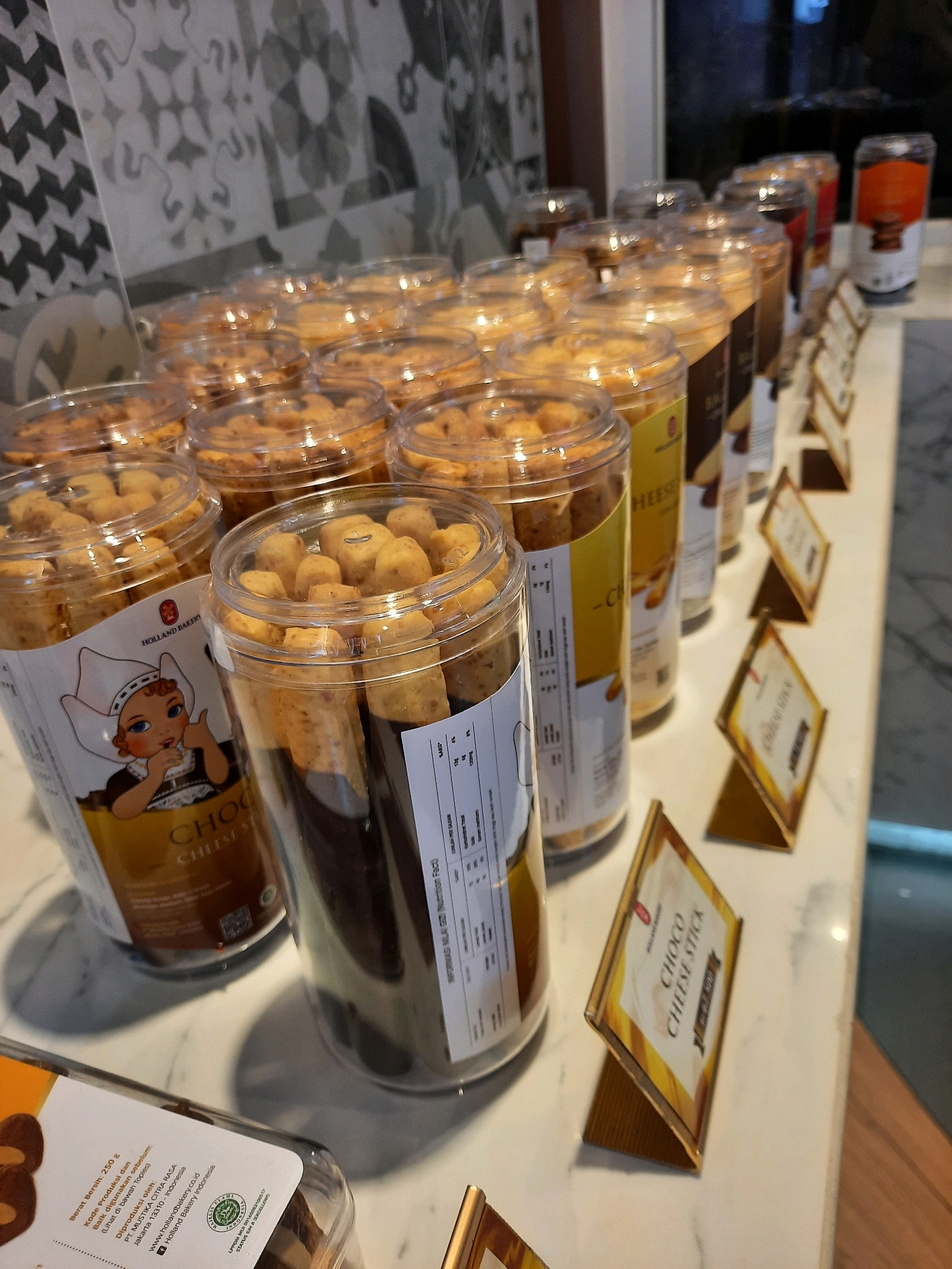 The image depicts a display of packaged baked goods, likely in a bakery or food stall located in East Jakarta, Indonesia. The foreground features several transparent plastic containers filled with cookies or crackers, arranged on a white countertop. One prominent container on the left shows a label with a cartoon image of a girl in traditional Dutch attire and text that includes "HOLLAND BAKERY" and "CHOCO CHEESE STICK". Another container to its right is also filled with similar snacks and has a label with nutritional information. In the background, rows of more packaged baked goods are visible, along with some display stands. The overall environment suggests a retail setting with a focus on food products.