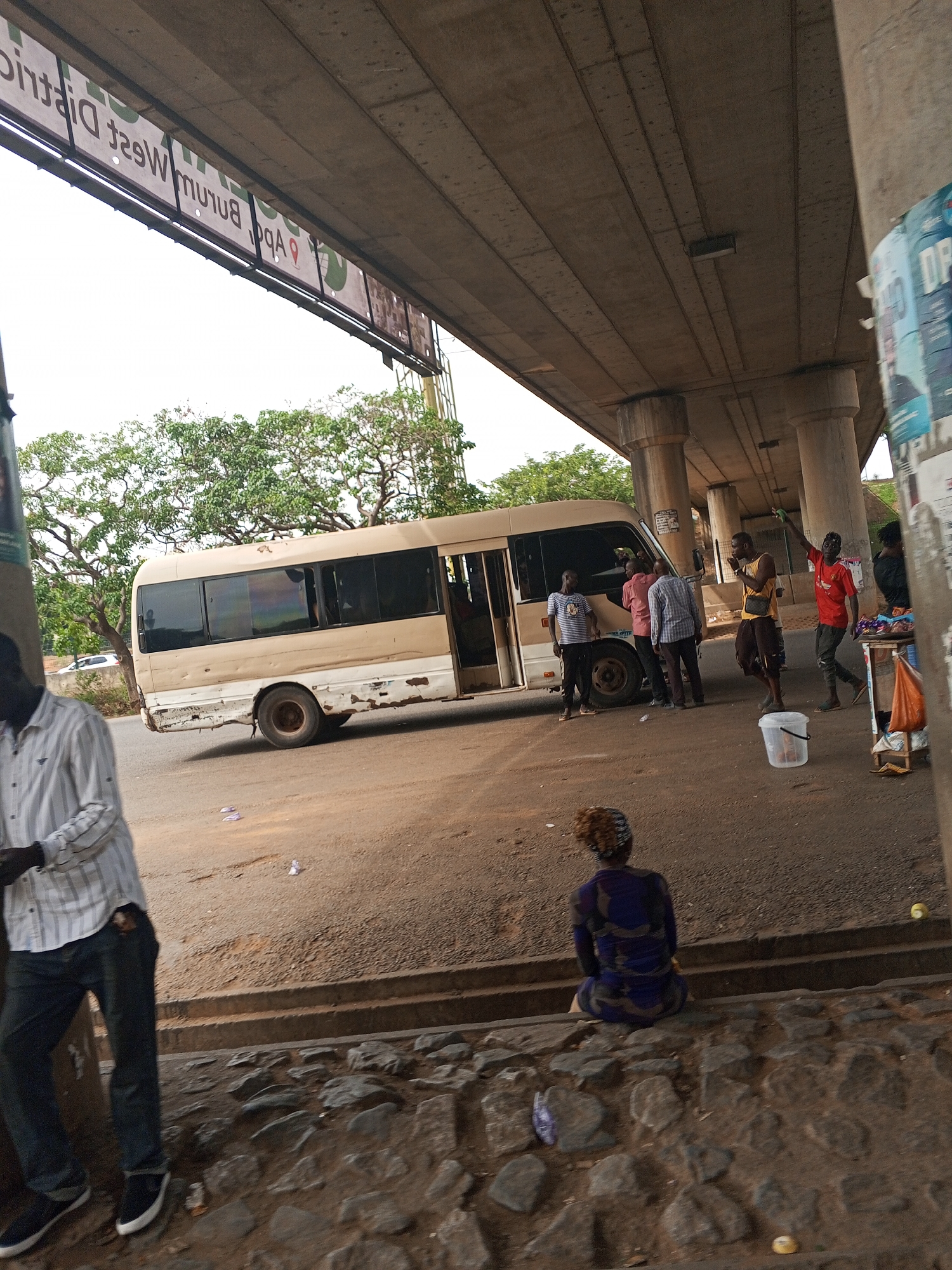The image depicts a street scene under an overpass in Abuja, Nigeria. A beige and white minibus with its door open is parked on the side of the road. Several people are standing near the bus, some appearing to be in conversation. Another person is seated on a low stone wall. A man stands near the left edge of the image wearing a striped shirt and jeans. A sign reading "Apo/Bumum West District" is visible attached to the overpass. The environment includes litter on the ground. A few vendors stand in front of a pillar selling food.