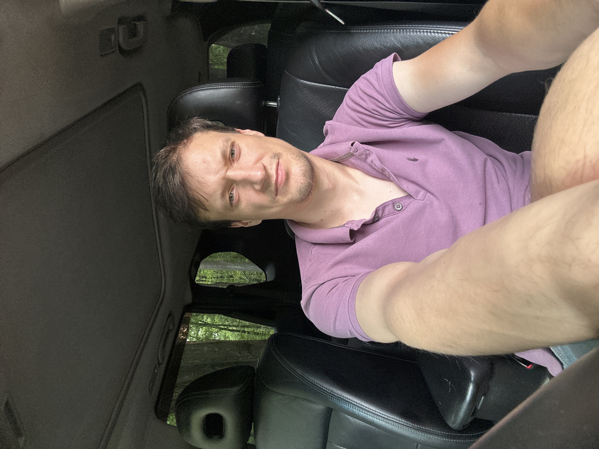 A man with short, dark hair and a light beard is seated in the driver's seat of a car. He is wearing a purple polo shirt. The car's interior is visible, with black leather seats and a sunroof. Outside the car, trees and foliage are visible through the windows, suggesting a natural, possibly wooded, environment. The lighting indicates it is daytime. There are no clear indications of the specific location within Shatava, Ukraine, or any specific actions beyond the man being in the car.