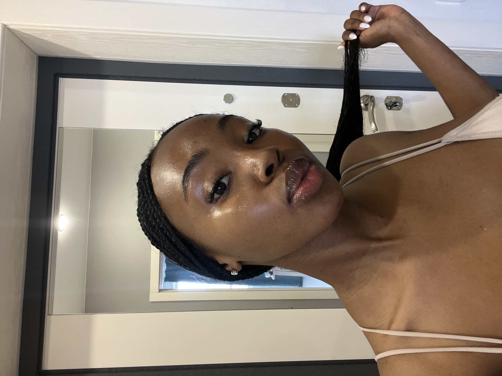 A young Black woman with braided hair is posing for a selfie in what appears to be an indoor setting. She is wearing a light-colored tank top. Her skin is glowing, and she is touching her hair with her right hand, which has manicured nails. In the background, a mirrored surface reflects the environment, showing a door with handles and a light fixture. The woman's expression is playful, with her lips slightly parted. The overall scene suggests a personal moment captured in a private space, possibly a home or hotel room. The image does not contain any information that would allow it to be located in the city of Kazan, Russia.