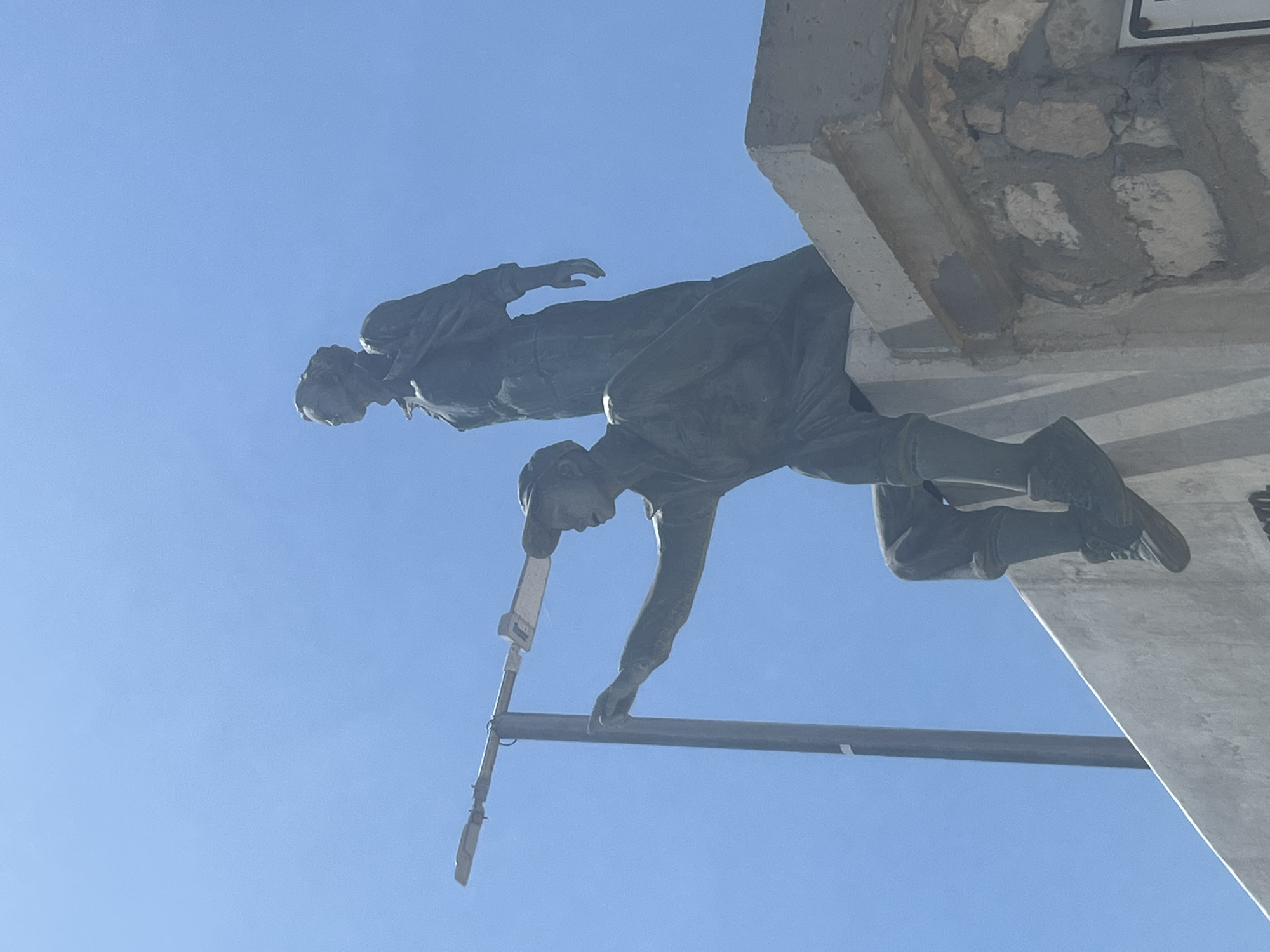 The image shows a bronze statue of two figures against a clear blue sky. The statue depicts a male figure in the foreground, seated on a concrete ledge and pointing forward with his right hand. He is wearing a cap, a shirt, and knee-high boots. Behind him, slightly taller, stands a female figure, also in bronze, looking to the left. The background is dominated by the bright blue sky, with a portion of a modern lamppost visible to the left of the male figure. The concrete ledge the statue sits on appears weathered and is partially made of stone. The statue is located in Varna, Bulgaria, specifically in the Sea Garden, commemorating the city's maritime history and the establishment of the Bulgarian Navy.
