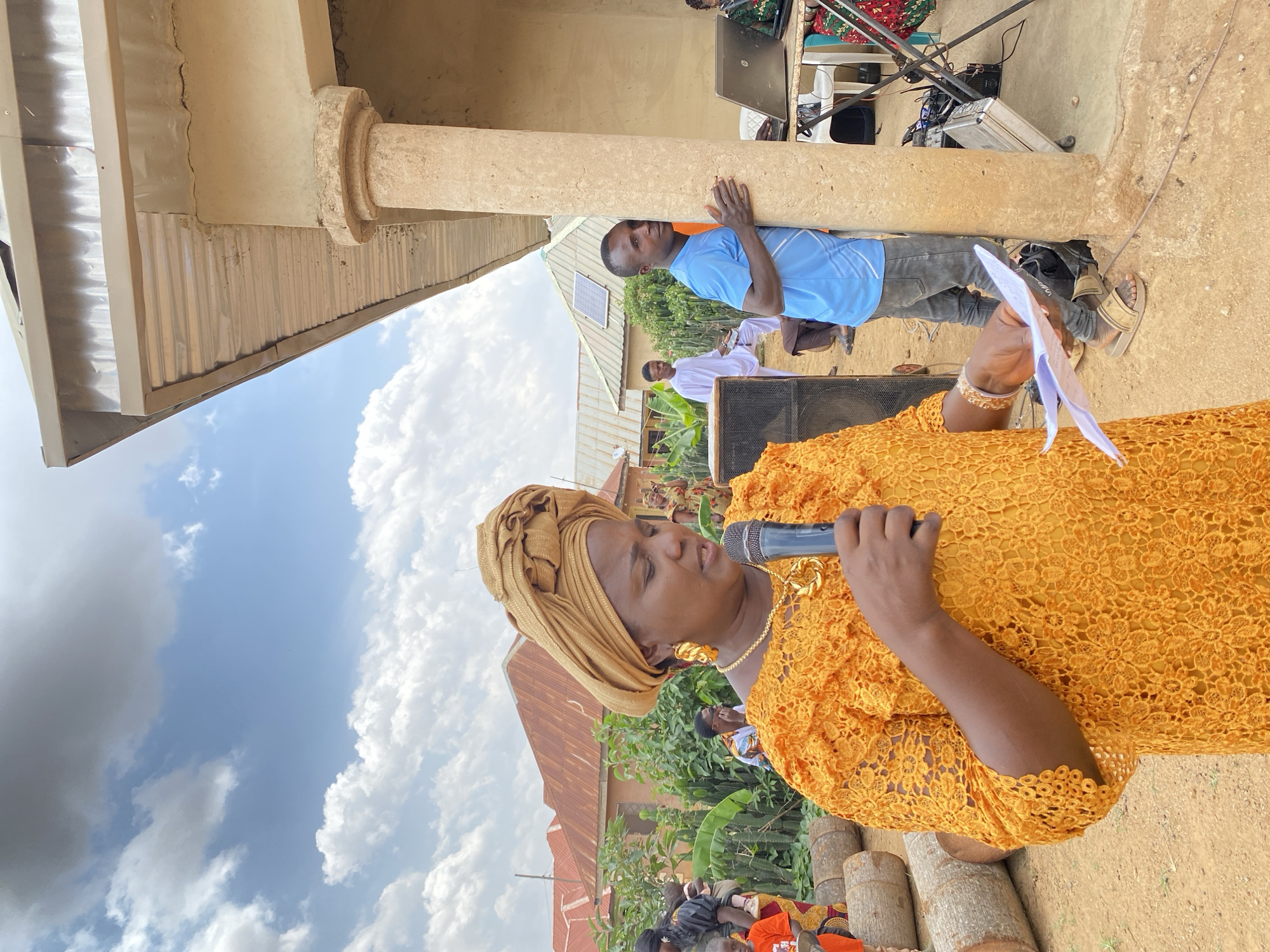 The image depicts a woman in a vibrant orange lace dress and a golden headwrap, speaking into a microphone while holding papers in her hand. She is wearing gold jewelry. Behind her, a man in a blue shirt is leaning against a column. In the background, there are people and buildings. The event appears to be taking place outdoors, in Barikin Sabon Gidan Kanat, Nigeria.