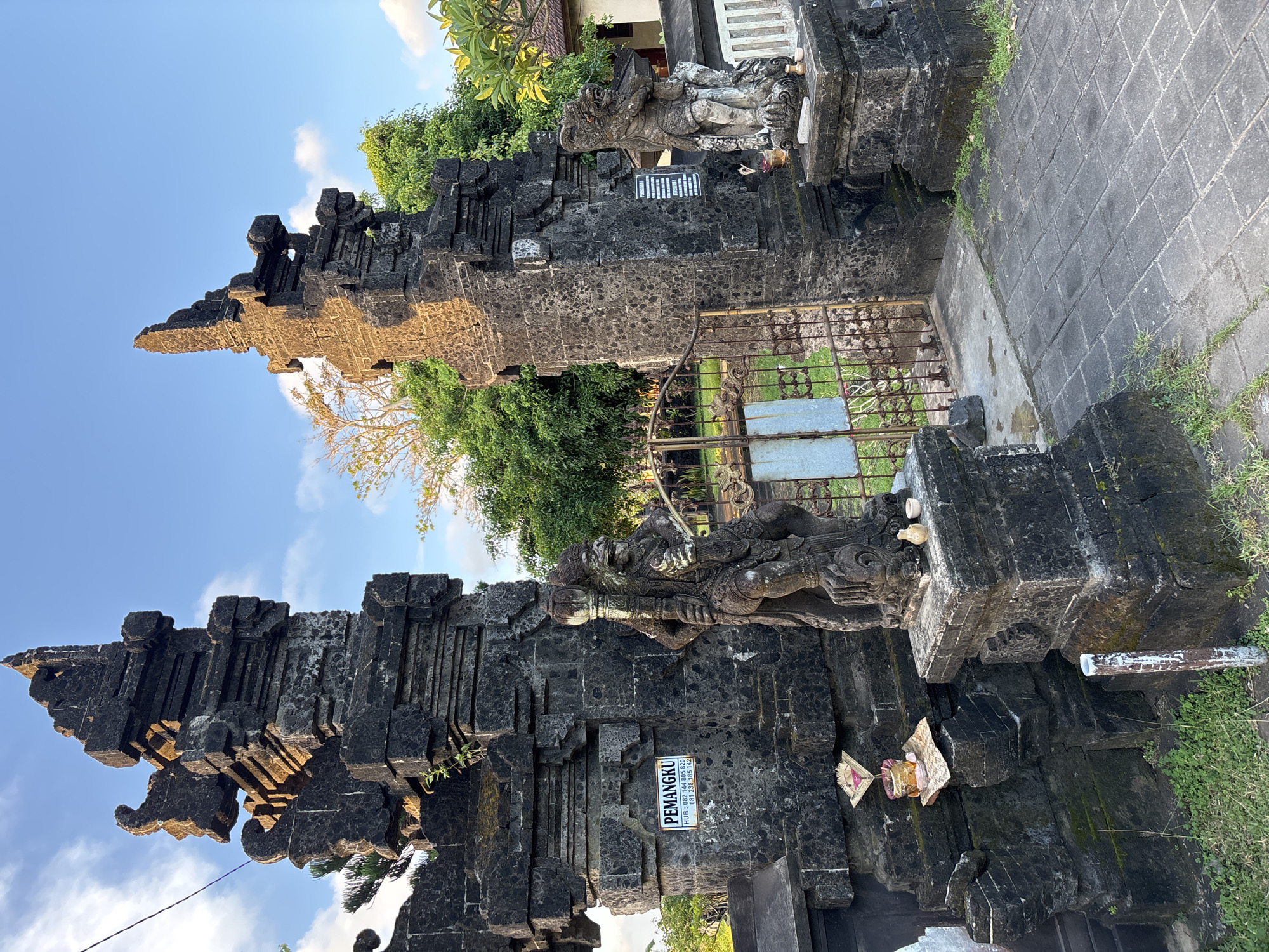 The image depicts a traditional Balinese gate in Kuta Selatan, Indonesia. The gate is constructed from dark grey stone, with a layered, pyramidal design. Ornate carvings and sculptures adorn the structure, including figures of deities or mythical creatures positioned atop the pillars. A metal gate is visible behind the stone structure, suggesting an entrance to a temple or sacred site. A small sign with the text "PEMANGKU" is attached to one of the pillars. The sky is blue with some clouds. Vegetation is present around the gate.