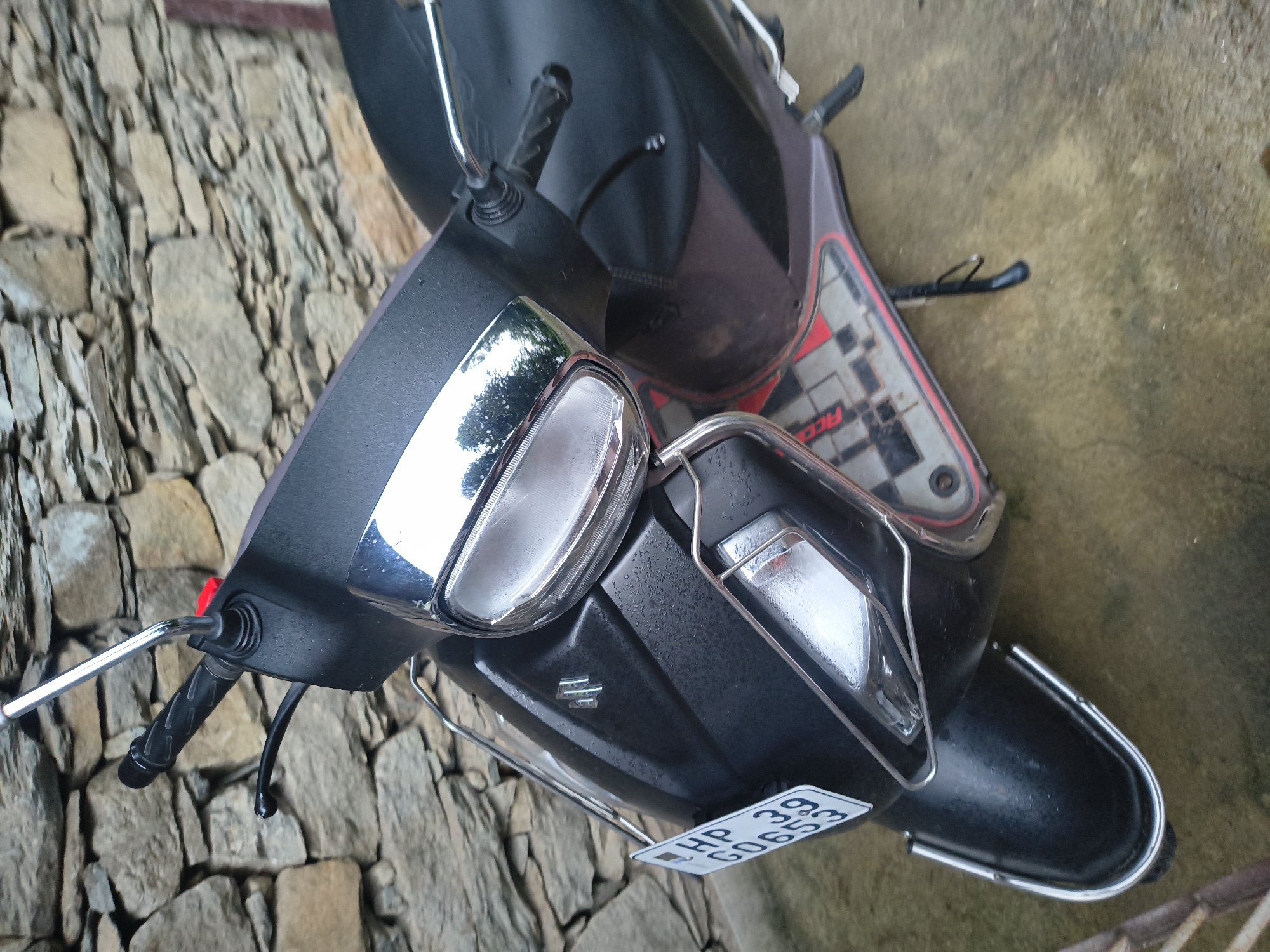 A black Suzuki scooter is parked on a wet, light-colored concrete surface. The scooter is positioned at an angle, with its front end facing towards the left and slightly upwards in the frame. The scooter's license plate, which reads "HP 39 G0653", is visible on the left side. The scooter has a chrome accent around its headlight and a chrome guard in front of the headlight. The background consists of a rustic stone wall. The scene appears to be an outdoor setting, possibly in Haini, India, judging by the license plate prefix. There are no people visible in the image. The scooter itself is slightly wet, suggesting it may have recently rained. The scooter has a dark colored seat and a patterned floorboard with red trim. A kickstand is deployed on the right side of the scooter.