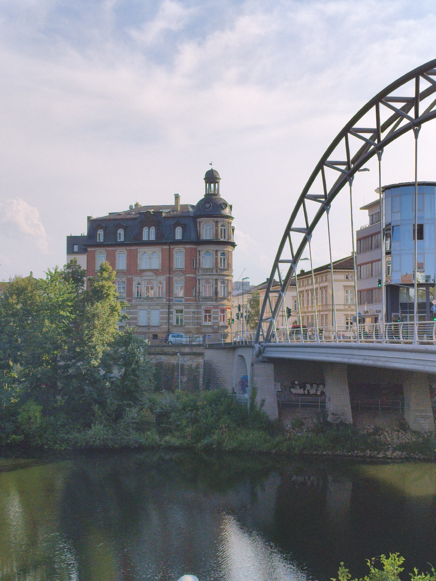 The image captures a view of the Regnitz River in Bamberg, Germany, with a bridge prominently featured. The bridge, with its distinctive arch, spans the river and carries a road with some traffic, including a light-colored car and traffic lights. On the left side of the bridge, a multi-story building with a red brick facade and an ornate mansard roof stands out. This building features numerous windows and architectural details. Adjacent to it, a cluster of lush green trees obscures a portion of the building and the riverbank.
On the right side of the bridge, a modern building with a glass facade reflects the sky and surrounding structures. The river itself is dark and reflects the bridge and the greenery along its banks. The sky above is partly cloudy, with soft white clouds scattered against a pale blue backdrop. The overall scene depicts an urban landscape where historical architecture meets modern design, bisected by the natural element of the river. The image is taken from a low angle, looking upwards towards the bridge and the buildings.