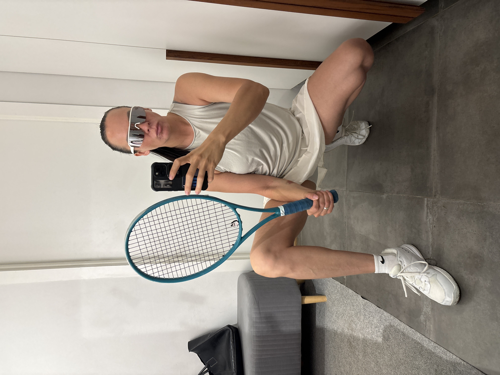 A person is squatting in what appears to be a dressing room or a similar enclosed space with a mirror. The person is holding a smartphone in one hand and a tennis racquet in the other, seemingly taking a selfie. They are wearing a white tank top, a white skirt or shorts, white socks with a black Nike logo, and white sneakers. They also have sunglasses on. The floor is a gray concrete. A padded stool is visible. A black tote bag is partially seen near the stool. Based on available information, a specific location in Chaikovsky, Russia cannot be determined from this image.
