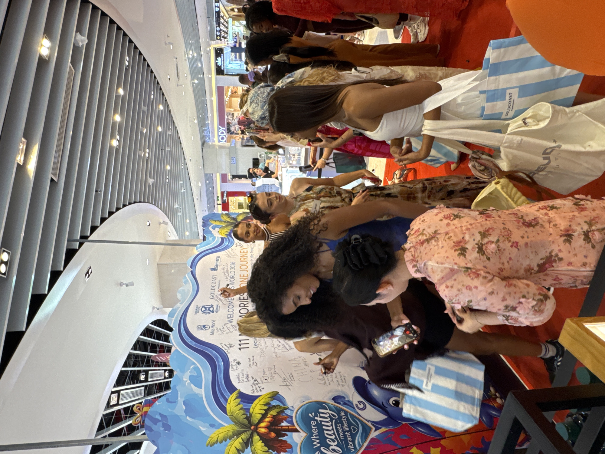 A group of young women, likely participants or staff in a beauty pageant or promotional event, are gathered in a shopping mall in Nha Trang, Vietnam. The women are diverse in ethnicity and are dressed in stylish, modern attire. Many are interacting with a large white wall that has been decorated with graphics of palm trees and blue waves, and is covered in handwritten signatures and messages. Some of the women are holding pens and writing on the wall, while others are posing for photos, with some using their phones. Several women are carrying tote bags, some of which are branded with "GOLDCOAST." The event appears to be taking place on a red carpet. In the background, there are signs for various shops, indicating a commercial setting. The overall atmosphere is festive and engaging.