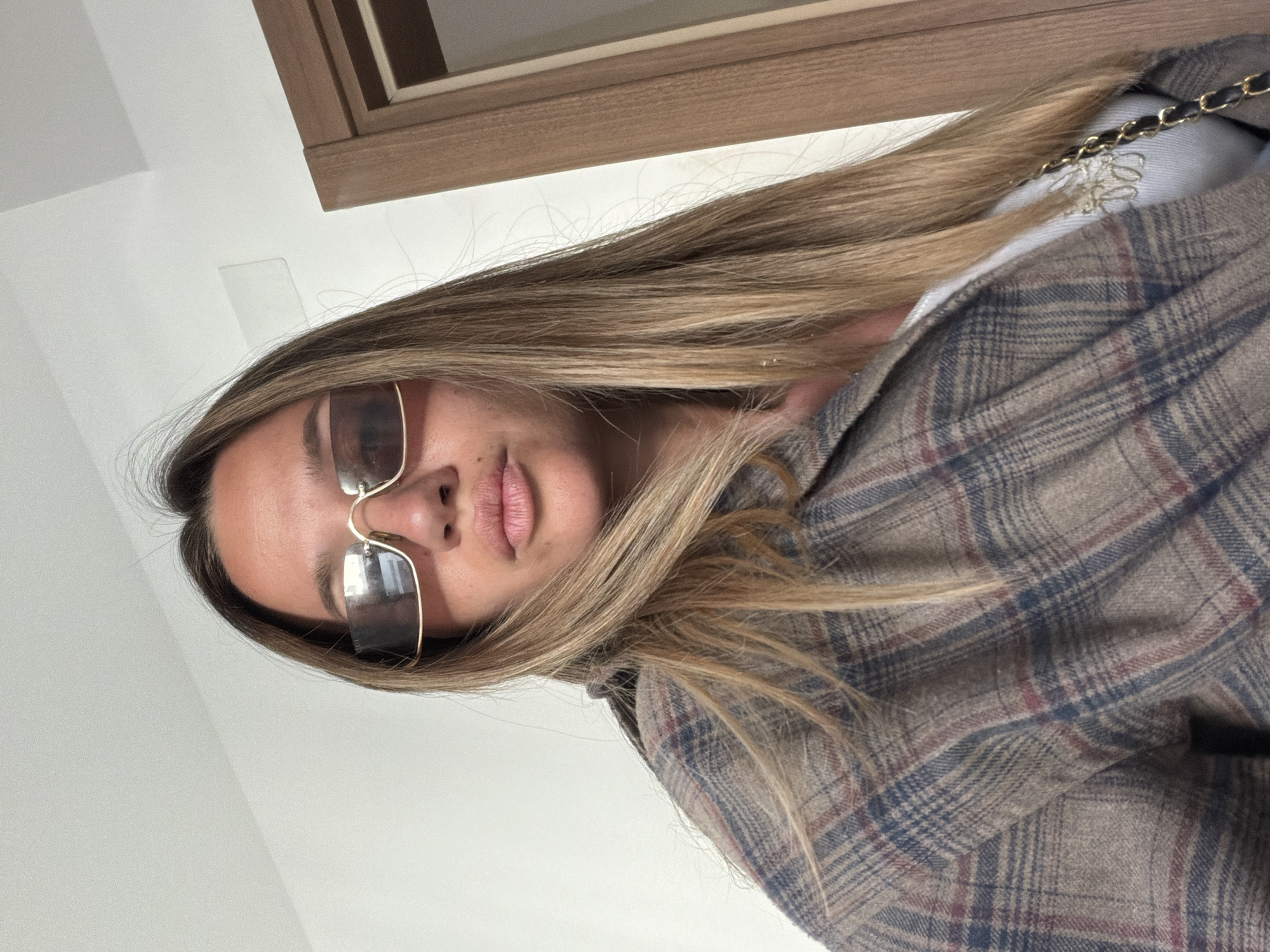 A woman is visible from the chest up, wearing sunglasses and a plaid jacket. A window is in the upper portion of the image. The location is Unknown, Unknown.