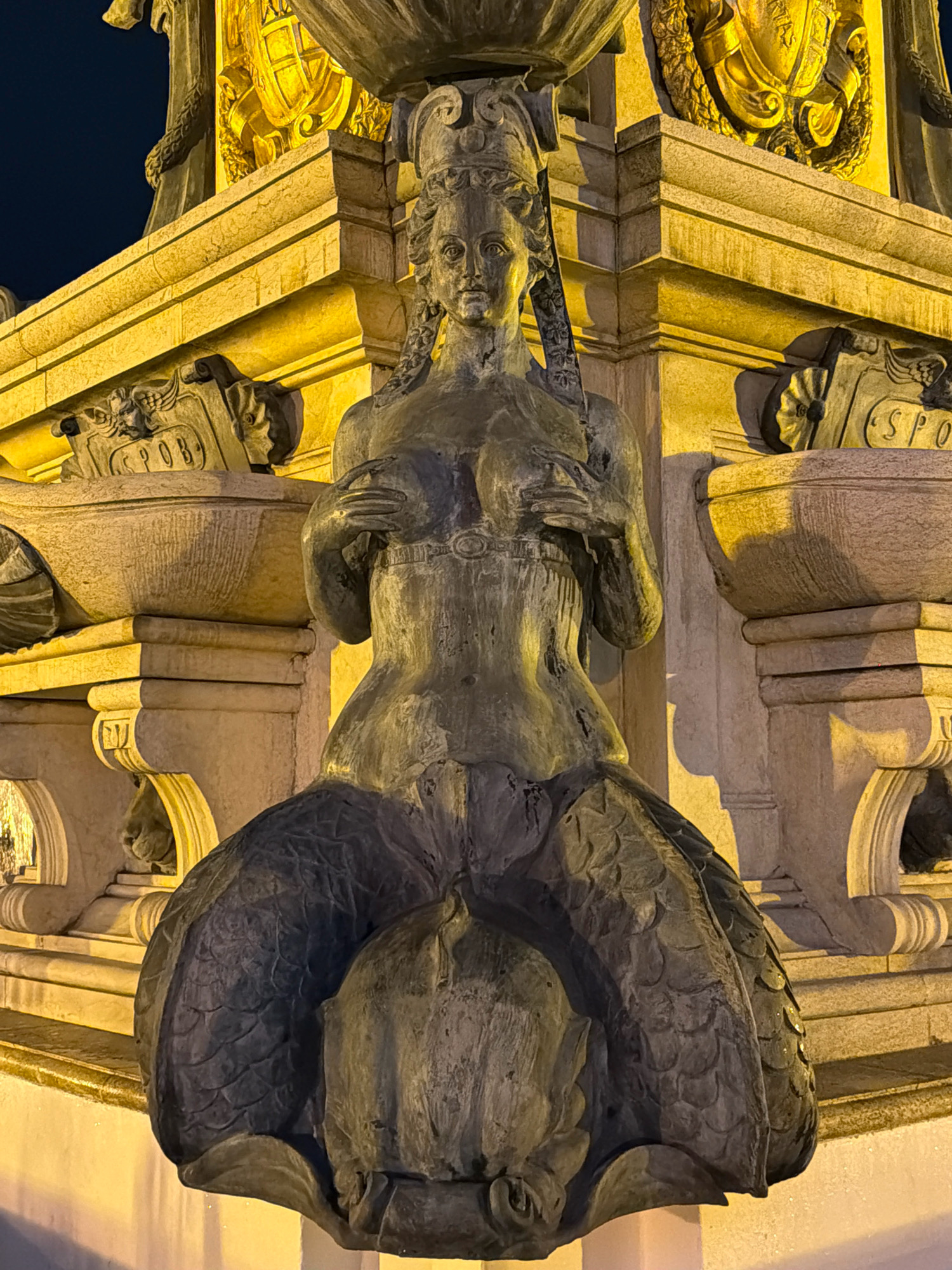 The image shows a bronze statue of a mermaid fountain located in Batumi, Georgia. The statue is positioned in the foreground, with its lower body transitioning into a fish-like tail covered in scales. The mermaid has her hands placed over her chest. Behind the statue, a large, ornate stone structure is visible, illuminated by yellow lights. This structure features carved details and what appear to be decorative bowls or basins. The background is dark, suggesting the image was taken at night.