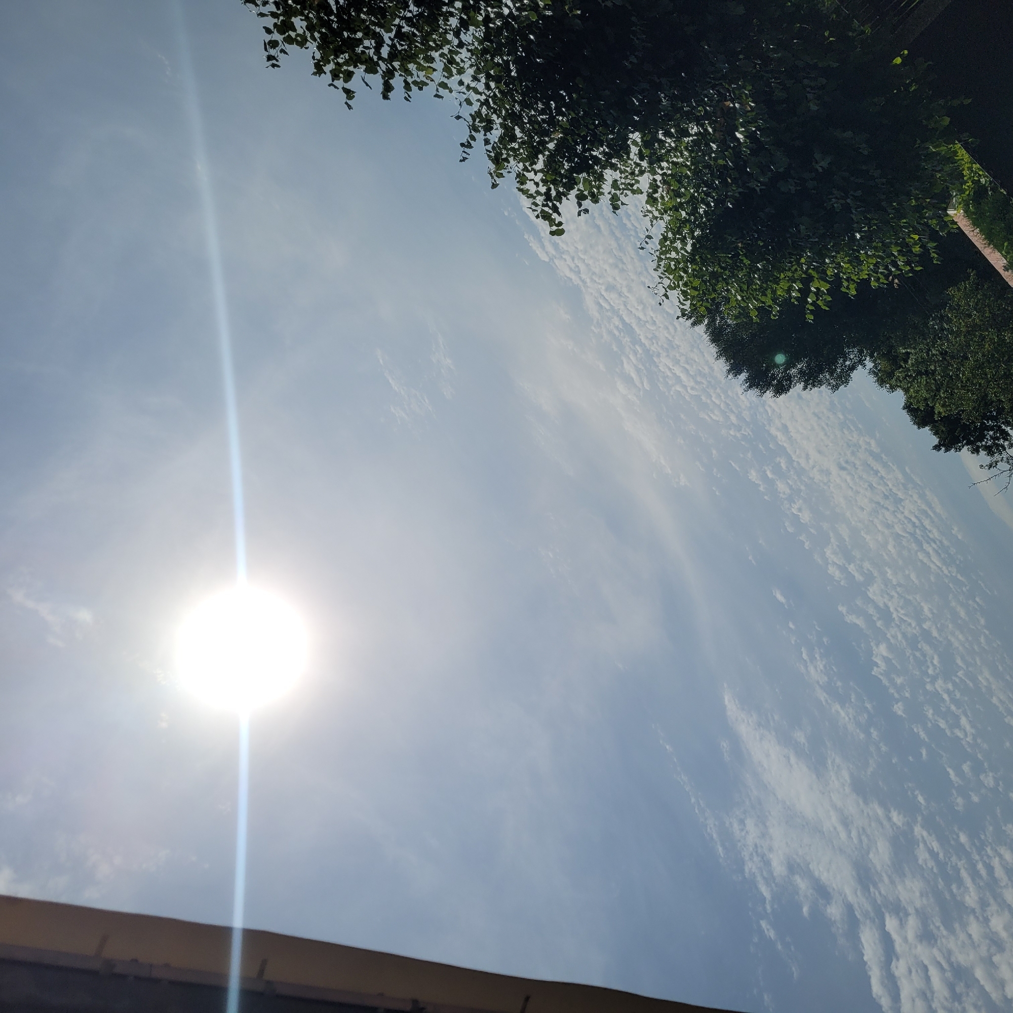 The image depicts a bright, sunny sky with scattered clouds over Jūrmala, Latvia. The sun is high and radiant, casting a lens flare across the sky. On the left side of the frame, a portion of a building's roofline is visible. The right side of the image is dominated by the lush green foliage of trees, with some leaves catching the sunlight. The overall scene suggests a clear day, likely during warmer months, in an outdoor setting within Jūrmala. No people or specific activities are discernible.