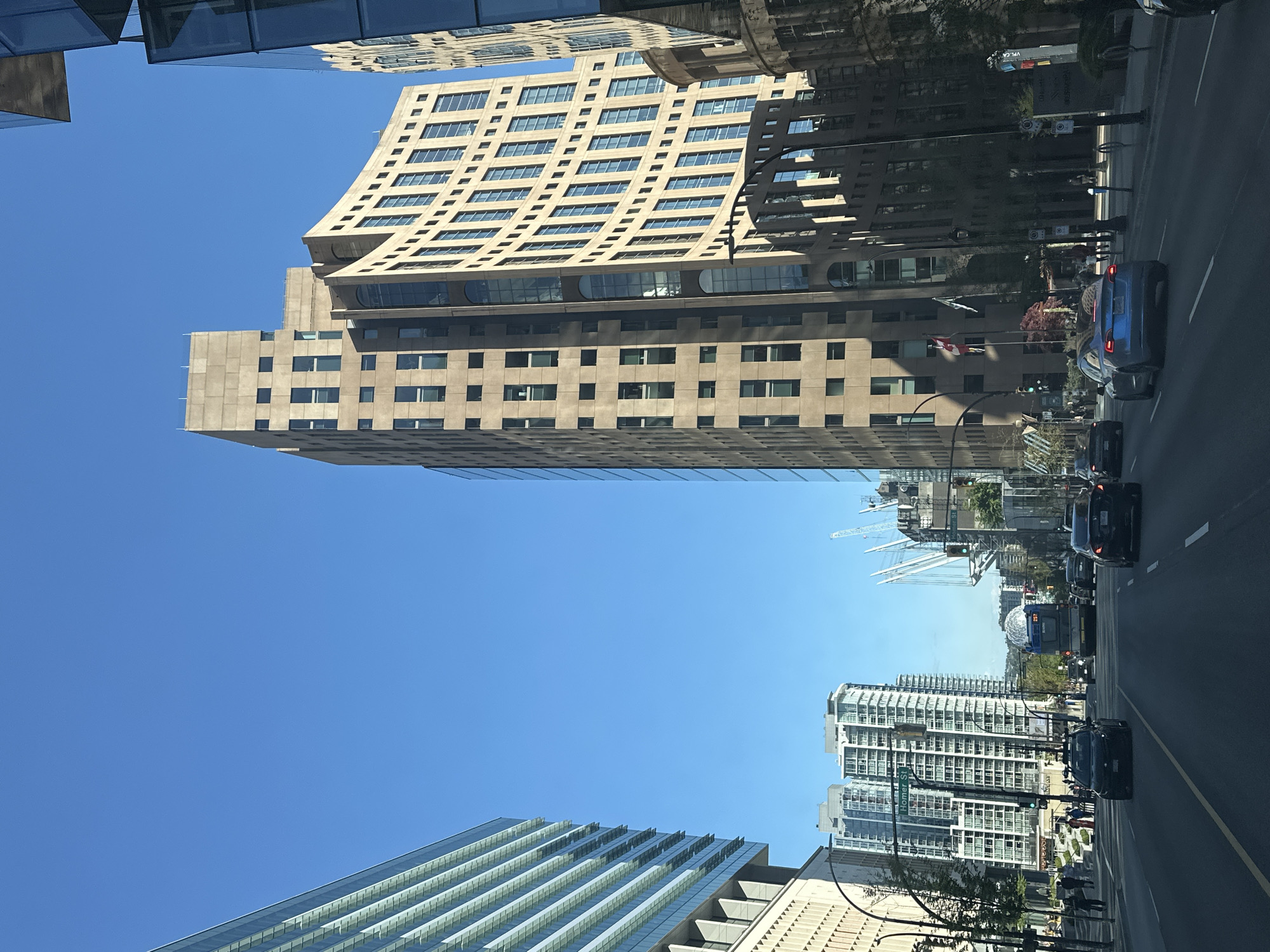 The image captures a street scene in Vancouver, Canada. The architecture is dominated by tall buildings, including the Fairmont Hotel Vancouver, characterized by its distinctive curved upper floors. Several cranes are visible in the background, indicative of ongoing construction. The street is populated with vehicular traffic, and a Canadian flag is noticeable, attached to one of the buildings. The sky is clear and blue.