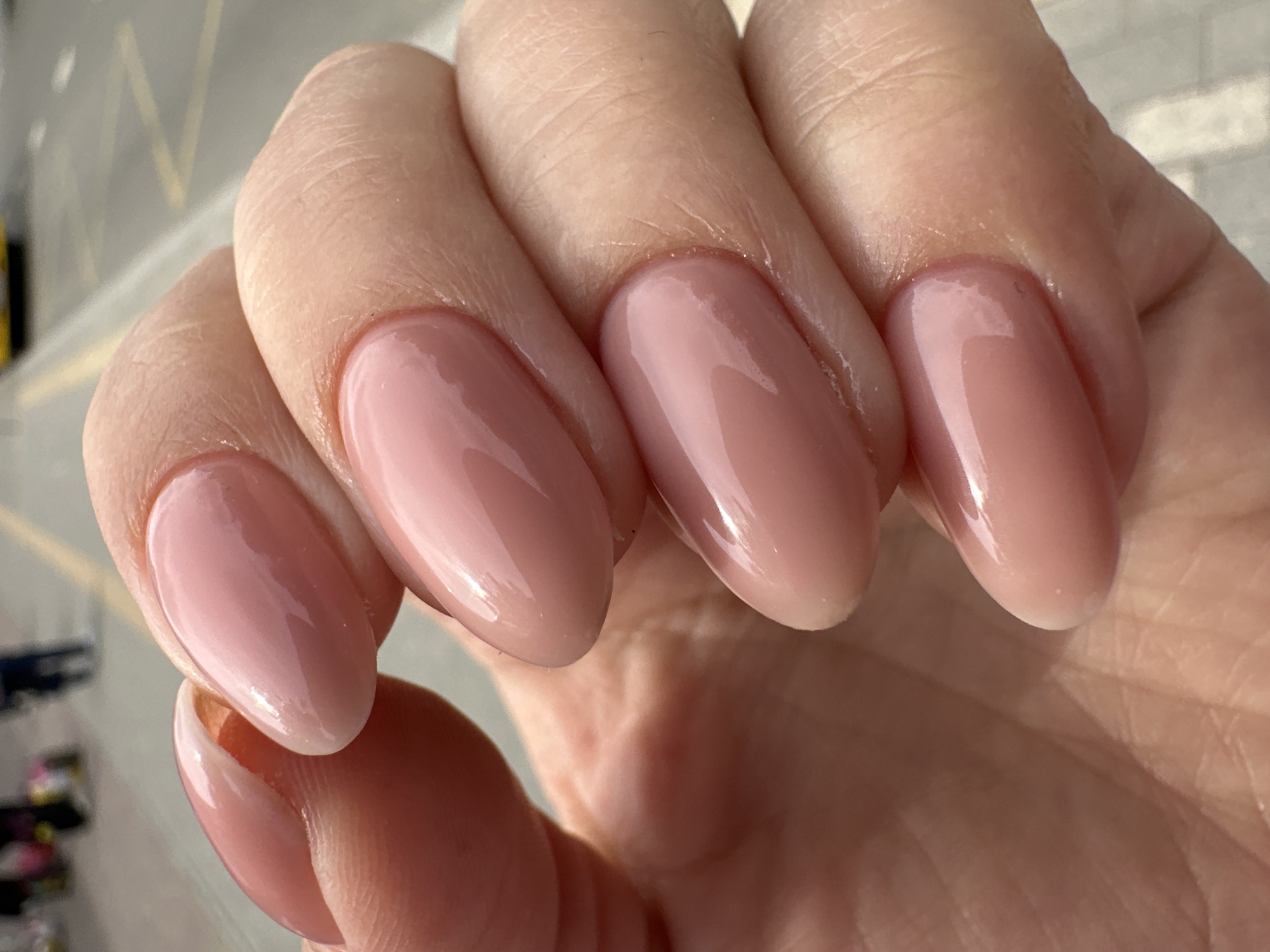 A close-up photograph captures a left hand showcasing five manicured nails, held against a brightly lit, blurred background. The hand belongs to an adult, with fingers slightly curled, presenting the nails prominently. Each nail is long, uniformly oval or almond-shaped, and coated in a high-gloss, translucent light pink or nude polish, giving a refined and natural appearance. The reflective surface of the polish shows distinct highlights from a strong, natural light source, indicating it is daytime.
The skin on the fingers displays natural texture, including subtle creases and fine lines, particularly noticeable around the cuticles and knuckles. The cuticle areas appear neat and well-cared for. The background is significantly out of focus, revealing abstract shapes and lines that suggest an outdoor urban environment, possibly a street or buildings, but no specific details or landmarks are discernible. There is no visible text in the image. The scene is presented without any specific activity or discernible emotion, focusing solely on the aesthetic of the hand and manicure. This image is contextualized as being from Ternopil, Ukraine.
