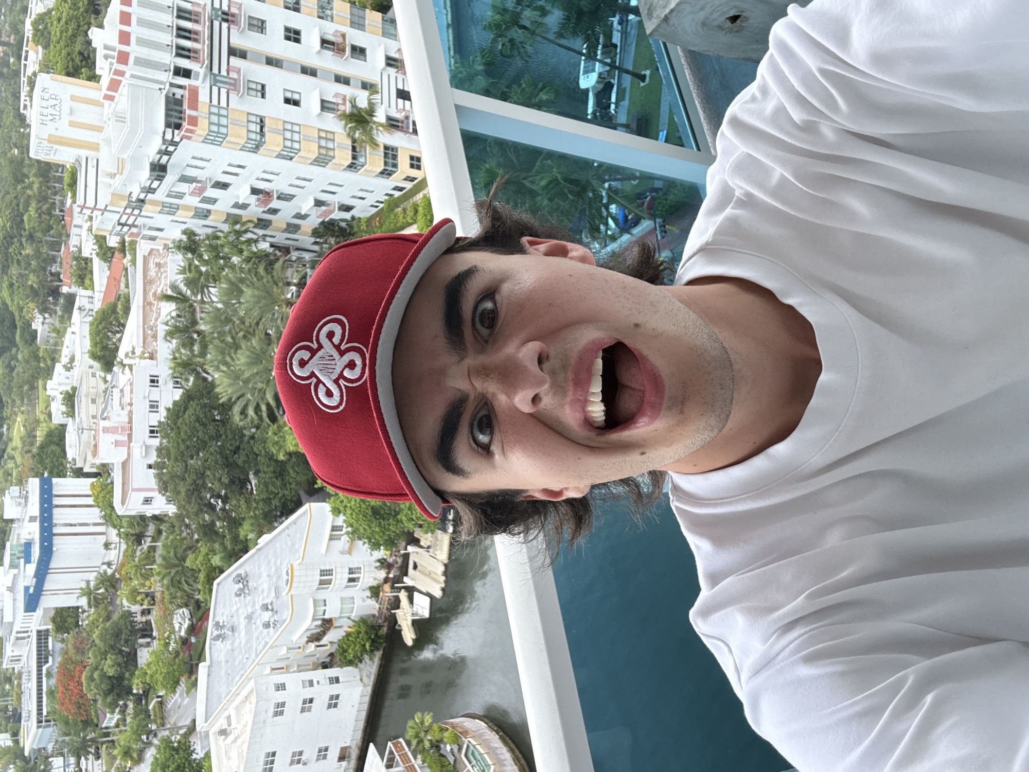 A person is taking a selfie on a balcony in Miami Beach, Florida, United States. The person is wearing a red baseball cap with a white clover-like design, and a white t-shirt. The person has dark hair and is making a funny face with their mouth open. The backdrop shows a waterway and buildings, and the “Helen Mar” building is on the left.