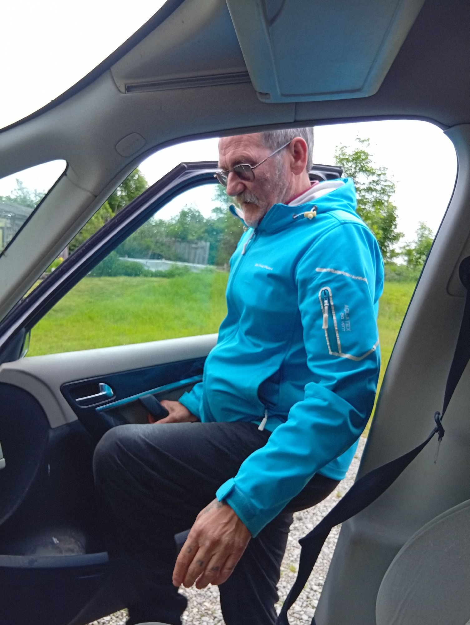 In Krikliai, Lithuania, a man with glasses and a gray beard, wearing a blue jacket, is entering a car. The car door is open, and the man's left leg is inside the vehicle, while his right leg is outside, with his foot on the ground. He is holding onto the car door. The interior of the car is visible, including the door panel and a seatbelt. Outside the car, there is a grassy area with some trees and a fence in the background, suggesting a rural or suburban setting. The weather appears overcast.