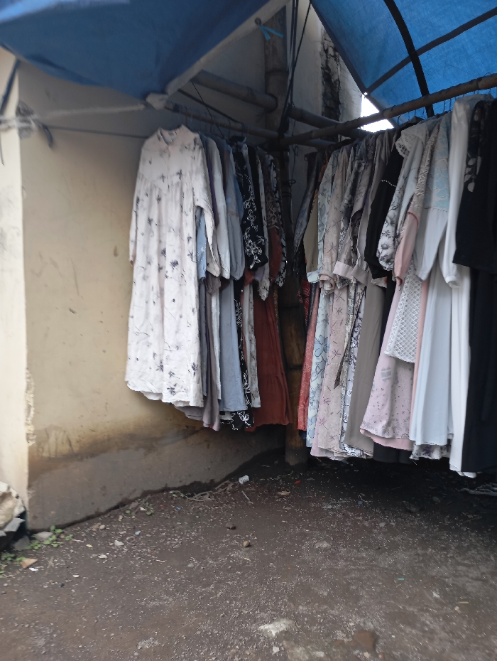 The image shows a row of clothing hanging from a metal pole under a blue tarp. The clothing is mostly women's dresses and tops, and they are all in various patterns and colors. The pole is attached to a concrete wall and a pillar. The scene appears to be in a narrow outdoor space, likely an alley or a market stall, with dirt floor and a concrete curb. The scene is located in Kutawaringin, Indonesia, at the coordinates -6.974, 107.519.