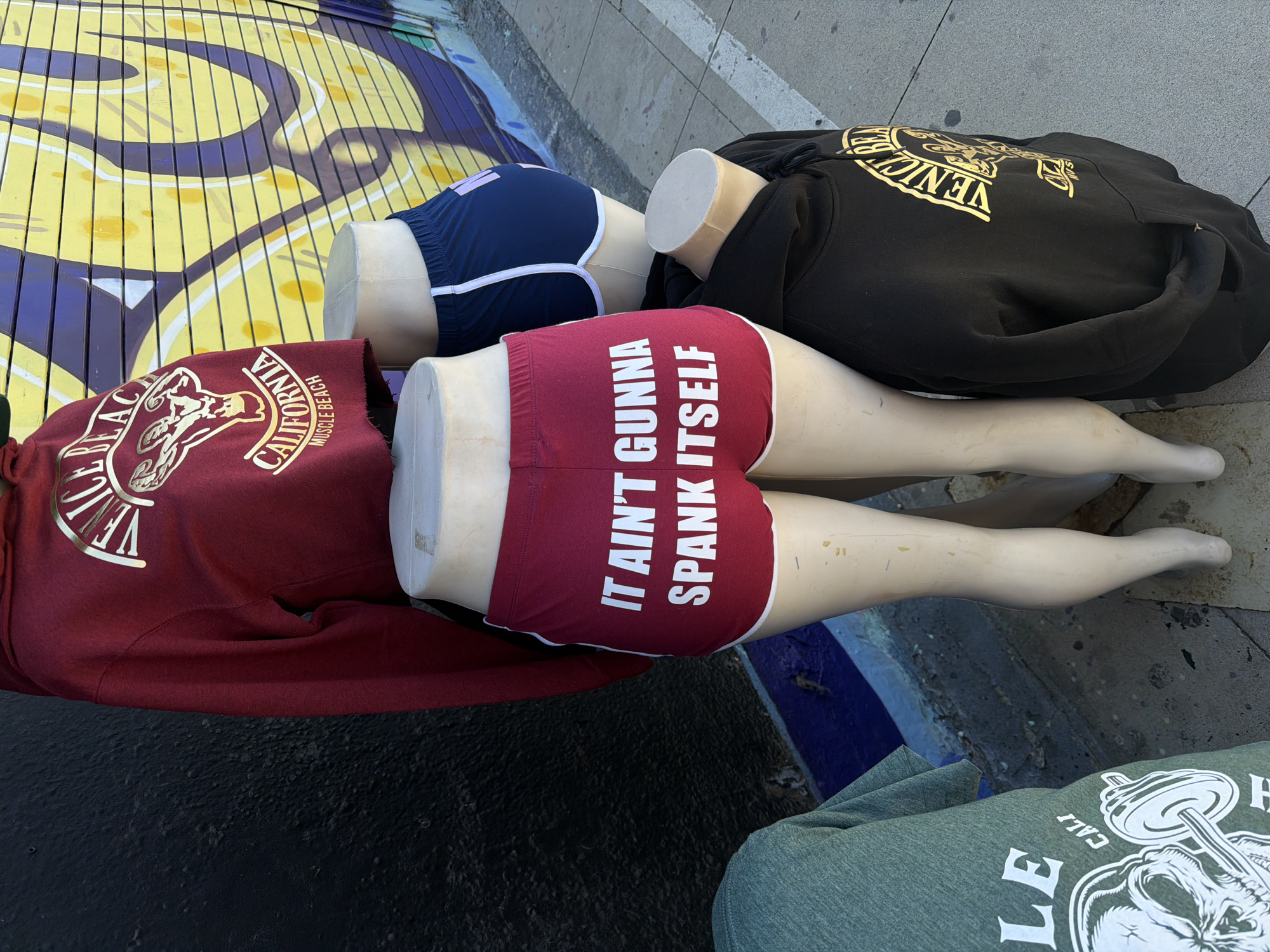The image displays a street-level clothing display featuring three partial mannequins laid on their sides on a concrete or paved surface. Behind them, a brightly painted wall or structure with a yellow and purple wave-like pattern is visible.
In the foreground, a mannequin wears a dark maroon hoodie with a gold-colored logo and text, which reads "VENICE BEACH CALIFORNIA MUSCLE BEACH." Below this, the mannequin wears red shorts with white trim, boldly printed with the white text "IT AIN'T GUNNA SPANK ITSELF." Its bare, pale legs extend forward.
Adjacent to this, another mannequin is dressed in a black hoodie with a gold "VENICE" logo. Part of a blue short with white trim is visible on its lower body. The third mannequin's legs are partially visible in the mid-ground, and a green t-shirt with a white logo featuring a barbell and an animal's head, along with "CALI," is seen in the bottom right corner.
The scene depicts merchandise, likely from a vendor or shop in Venice Beach, Los Angeles, United States of America, given the branding on the apparel. The overall lighting suggests it is daytime.