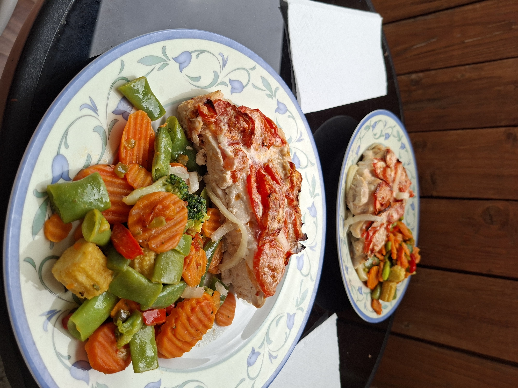 The image depicts two plates of food on a tray, possibly outdoors on a wooden surface. The plates have a similar design with a light-colored background and a blue floral pattern around the rim. Each plate contains a similar meal: a portion of cooked meat topped with sliced tomatoes and onions, accompanied by a mix of sauteed vegetables, including sliced carrots, green bell peppers, broccoli, and what appears to be yellow squash. There are also two white napkins near the plates. The backdrop appears to be wooden planks or a fence. The location cannot be determined definitively from the image, but the style of the meal suggests a home-cooked or casual restaurant setting. The location of Tallinn, Estonia, cannot be confirmed from the image alone.