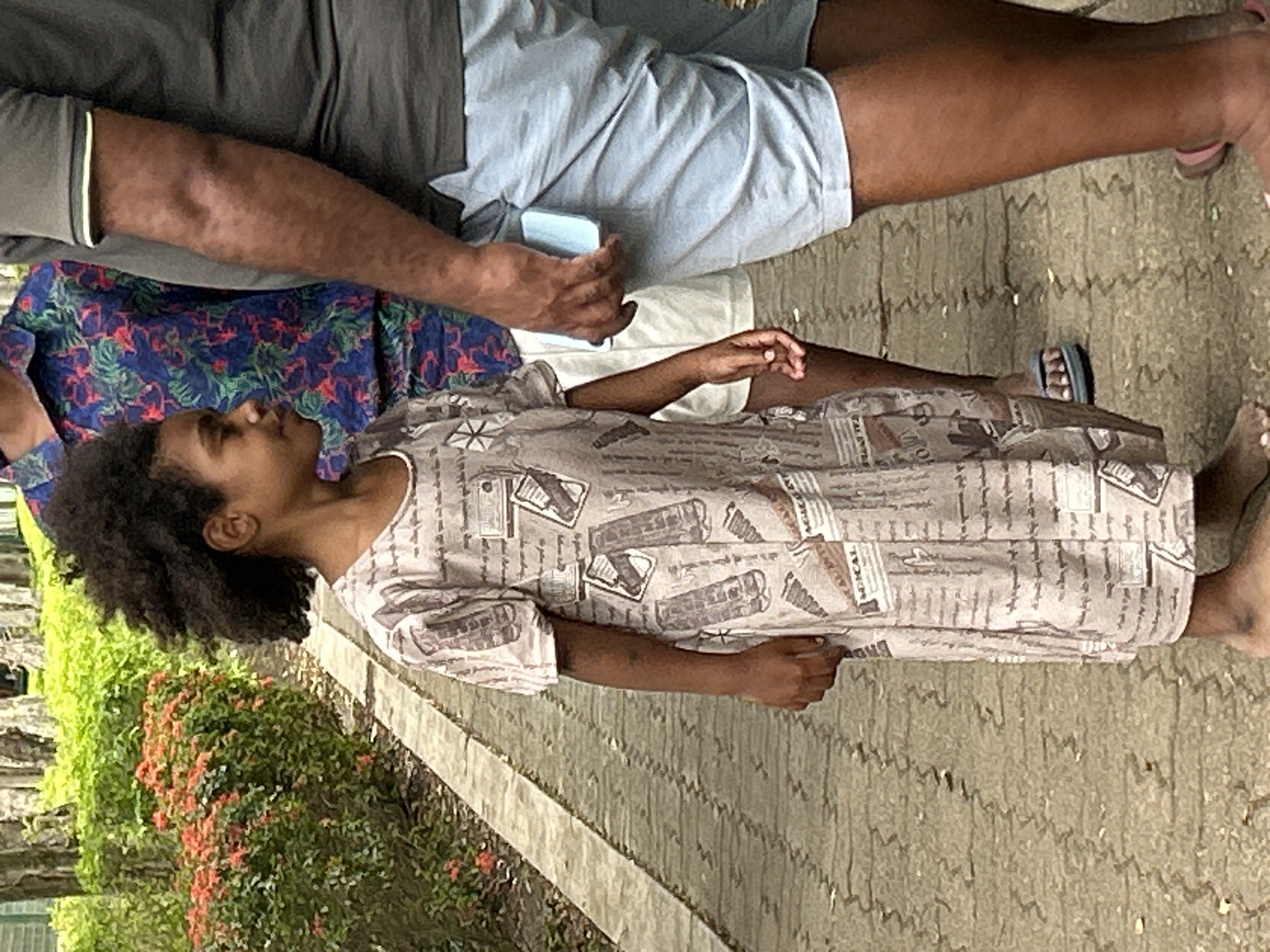 In Tanubada, Papua New Guinea, on a paved walkway lined with lush greenery and flowering bushes, three individuals are visible. A young person, standing on the left, wears a long, patterned dress with text and images resembling documents. To their right, a person in a floral shirt stands, partially obscured. On the far right, an adult, viewed from the waist down, wears a grey polo shirt and light blue shorts, holding a light blue object, possibly a phone. The environment suggests a park or garden setting with trees and foliage.