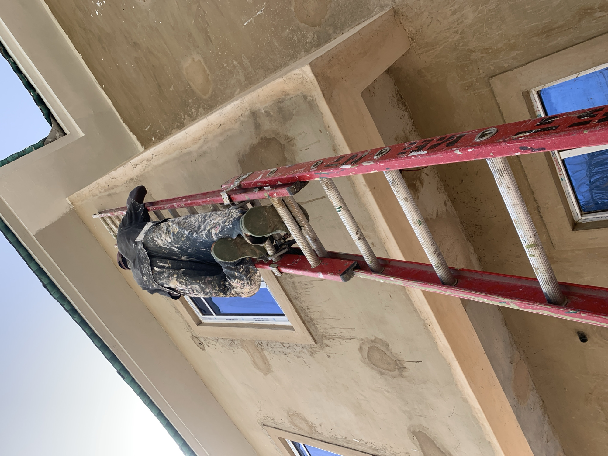 In Bukuru, Nigeria, a person wearing camouflage pants and a dark jacket is climbing a red A-frame ladder up the side of a building under construction. The building has a light brown stucco exterior with visible imperfections, and green trim along the roofline. Two windows with blue reflective glass are visible on the building's exterior. The sky is clear and bright.