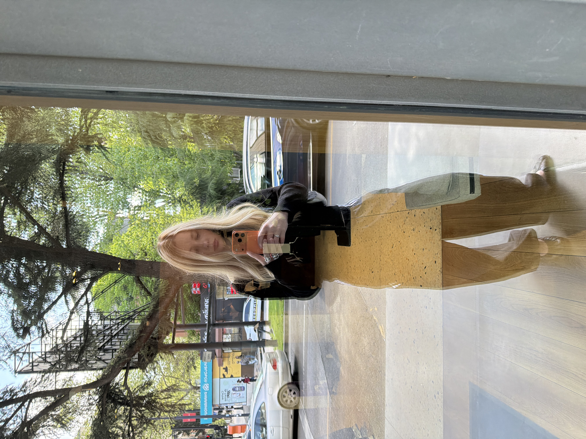 A mirrored image captured through a window reflects a woman holding a phone. She is wearing a black top. Outside the window, the reflection reveals a street scene with trees, buildings, billboards, and moving cars. The signs display text in what appears to be Georgian script, with the address "D. Uznadze Street, 2" visible on a blue and white sign. The surroundings indicate the location is likely Tbilisi, Georgia. The interior features wooden flooring and a light-colored rectangular structure, possibly a seating area or display.