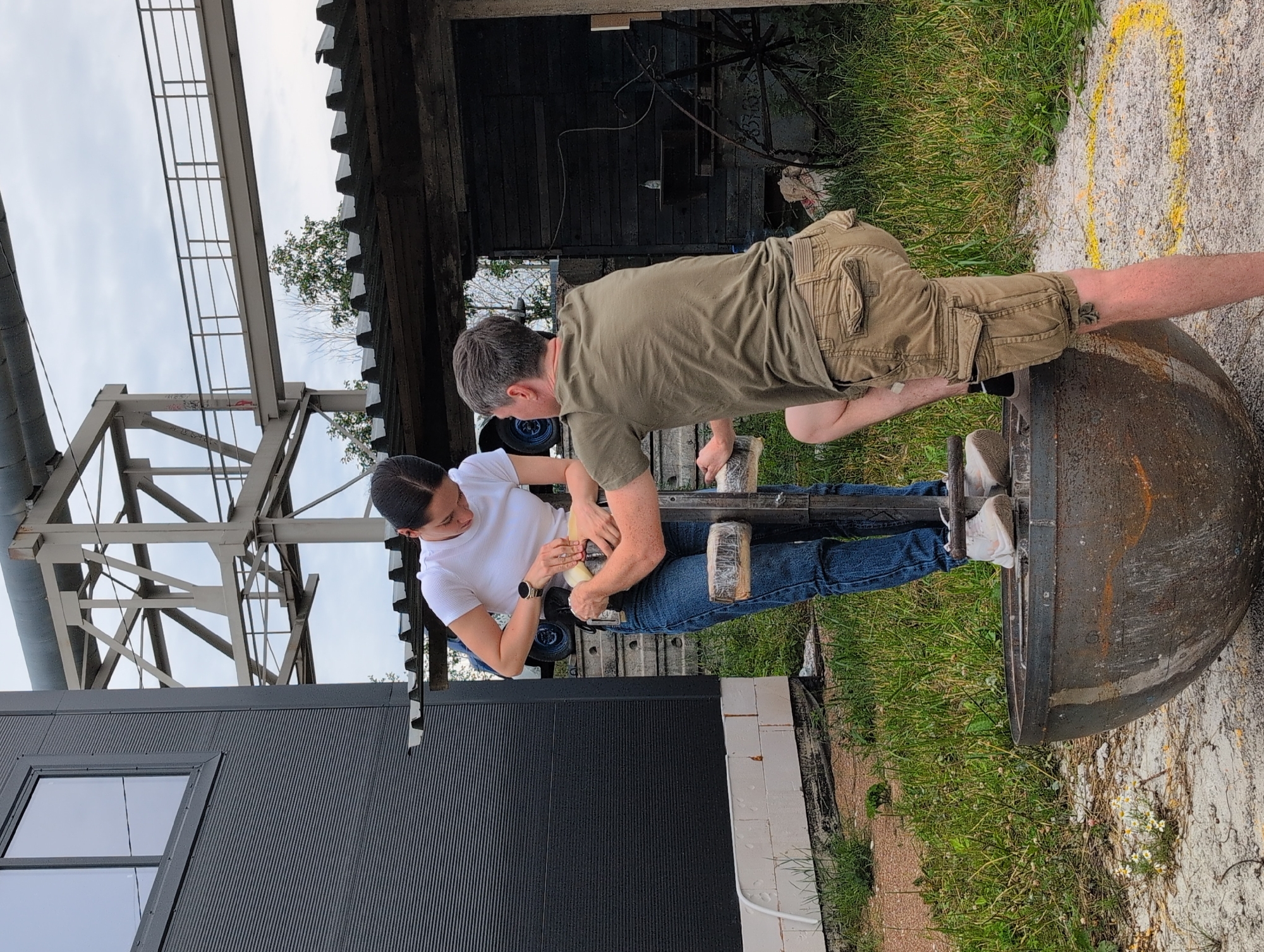 In Novoe Devyatkino, Russia, two individuals are engaged in an activity outdoors near industrial structures. A woman wearing a white t-shirt and blue jeans stands on the rim of a large, weathered metal basin, holding a tool and appearing to adjust something on a metal apparatus. A man, dressed in a green t-shirt and khaki cargo shorts, kneels beside the basin, his left leg extended, and assists the woman with the apparatus. The environment includes tall, grey industrial metalwork in the background, a dark-colored building with a window on the left, and a rustic wooden structure with a corrugated roof. Grassy vegetation and sand cover the ground, with a yellow painted circle visible on the ground near the man. The overall scene suggests an outdoor workshop or industrial area.