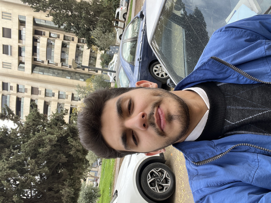 The image displays a headshot selfie of a young adult male, appearing to be in his late teens or early twenties, situated in the Kafr Sousa Municipality, Damascus, Syria. The male, positioned centrally in the foreground, has dark hair, dark eyes, and a short beard and mustache. He is wearing a white collared shirt beneath a black V-neck sweater with a diamond pattern, covered by a blue zippered jacket. He maintains direct eye contact with the camera and exhibits a slight smile with visible dimples on his cheeks.
Behind the individual, an urban street scene unfolds under what appears to be an overcast sky. To the left, a white modern SUV is partially visible, showing its front wheel and a section of its body. To the right, the rear section of a dark blue sedan is visible, with its driver's side window showing scattered debris. Further back, additional vehicles are parked along a street. The background features a prominent, tall, multi-story building characterized by a light-colored facade, numerous balconies, and expansive glass windows, indicative of modern architectural design. Green trees with dense foliage and shrubs are present throughout the background, bordering the street and the building. A modern streetlamp with multiple fixtures is visible between the parked cars and the background building.