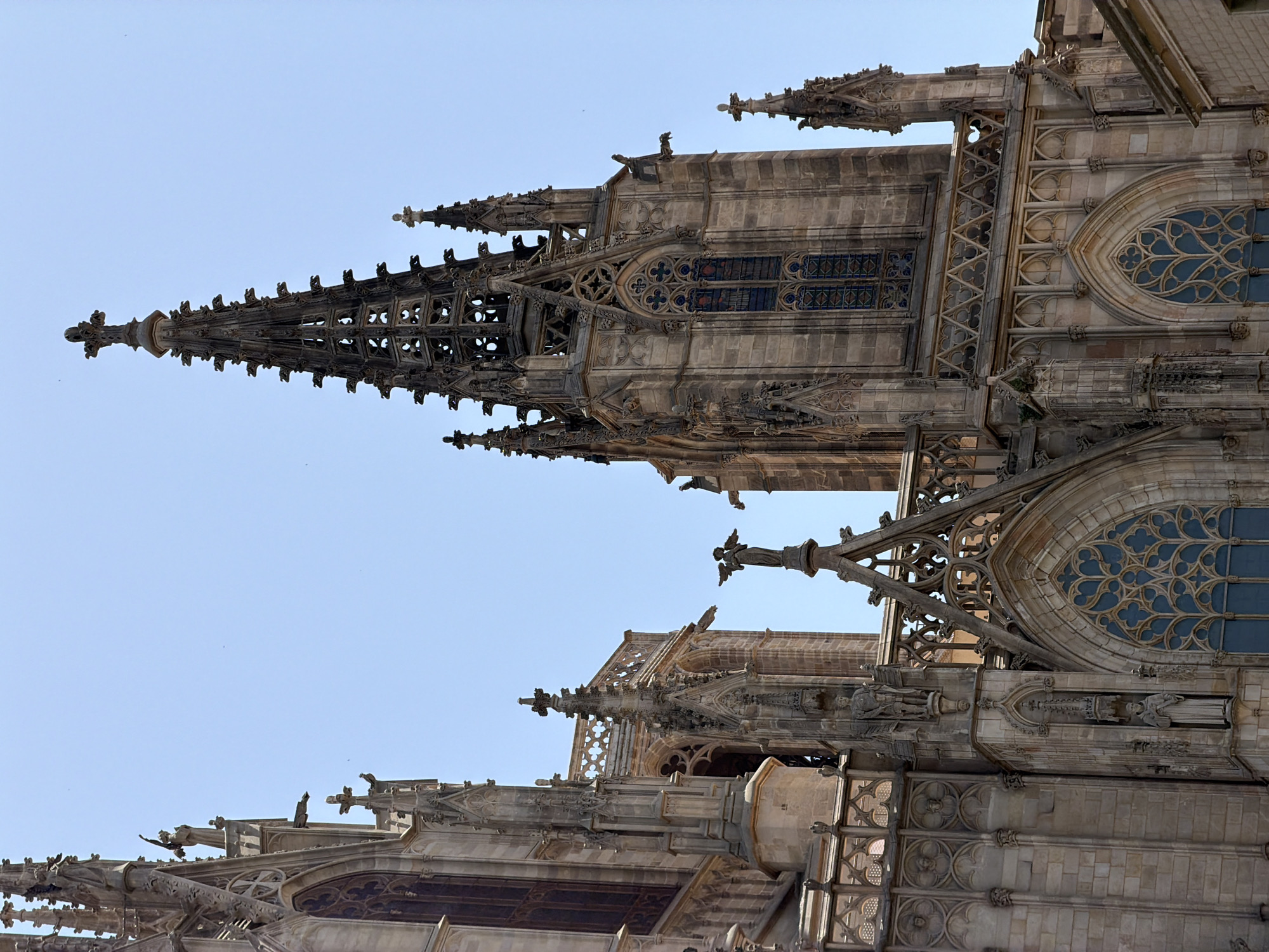 The image shows a close-up of the exterior architecture of the Barcelona Cathedral, located in the Gothic Quarter of Barcelona, Spain. The detailed Gothic design features intricate stonework, spires, statues, and stained-glass windows against a clear blue sky. The architecture is characterized by pointed arches, elaborate tracery, and ornate embellishments. Several statues of figures, possibly saints or angels, are visible on the facades and spires. Gargoyles also adorn the building, typical of Gothic cathedrals. The material appears to be weathered stone, suggesting age and historical significance. No people are visible in the immediate scene, and the focus is entirely on the majestic structure of the cathedral.