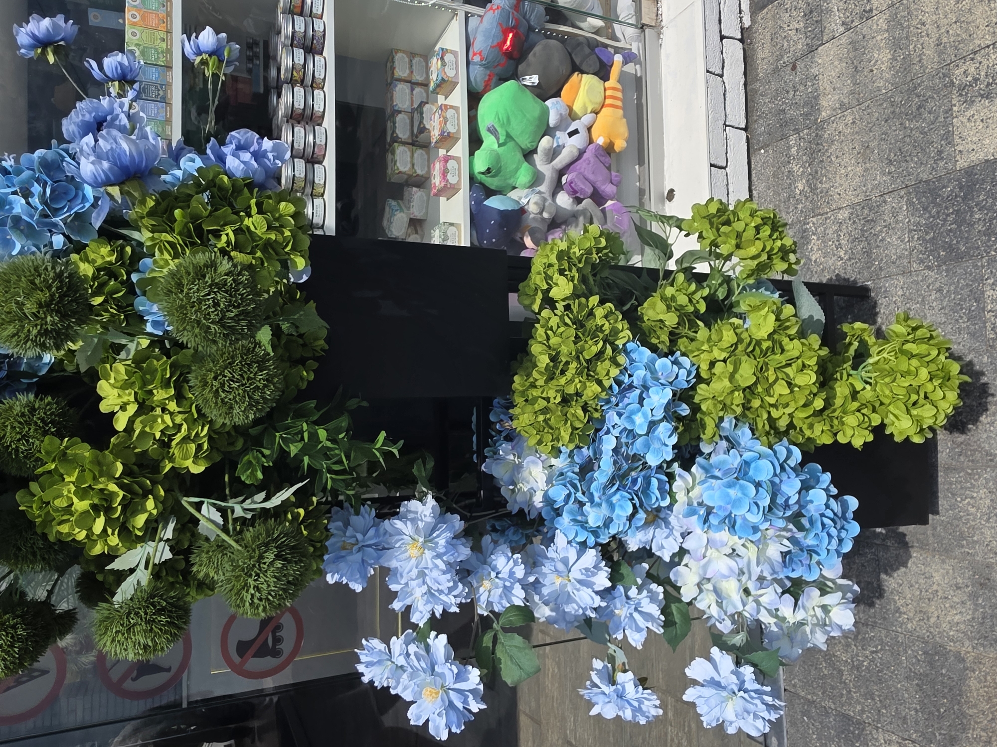 A storefront displays an arrangement of artificial flowers, primarily in shades of blue and green, in a black planter. Behind the flowers, a glass-fronted display case is visible, filled with shelves of small jars and boxes, and several plush toys including a green frog, a purple character, and an orange and yellow striped character. The display case is set against a white brick wall. The flowers are in the foreground, casting shadows on the grey tiled pavement. Signs are visible on the left side of the image, including a red circle with a diagonal line through a figure on a skateboard and another sign with a similar red circle and line through a person walking. The environment suggests an outdoor urban setting, potentially in Sochi, Russia, given the context.