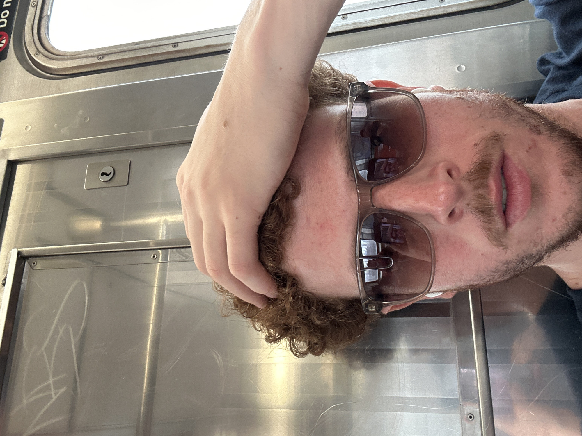 A selfie of a man on a New York City subway train. He is wearing sunglasses and has his hand on his head. The subway car is stainless steel and has graffiti on the window. The train is likely in New York City, as indicated by the MTA signage.