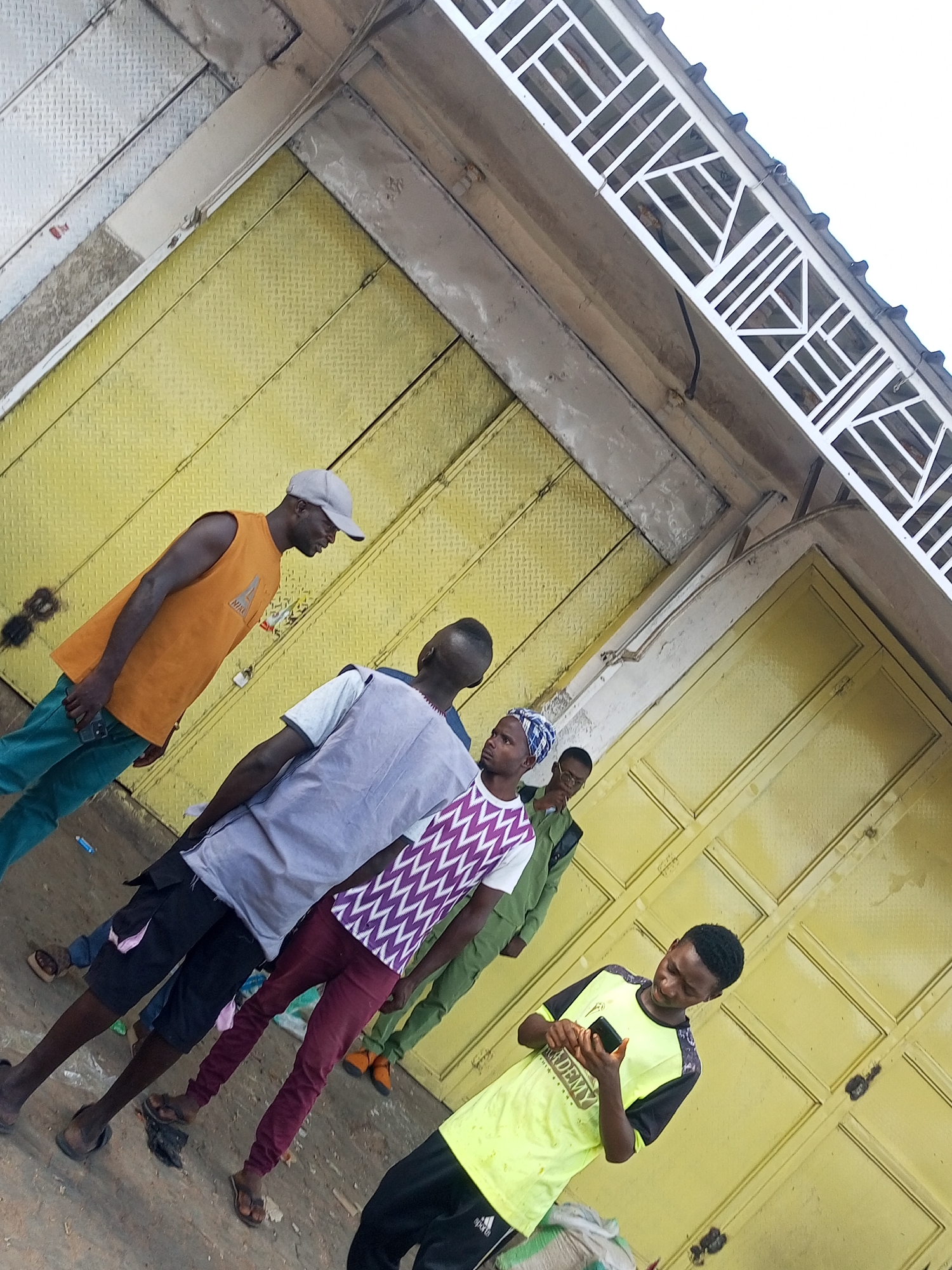 The image depicts a group of young men standing outside buildings with yellow metal doors. The perspective is from a high angle, looking down at the scene. In the foreground, a young man in a bright yellow t-shirt with black sleeves is looking down at his phone, which he is holding in both hands. To his left and slightly behind him, another young man in a patterned purple and white shirt and maroon pants stands facing away from the camera, looking towards his left. Further to the left, a man in a light gray shirt and black shorts is also looking away. Another man, wearing an orange sleeveless shirt and teal pants, stands to the left of the man in gray, also facing away. The background consists of the yellow metal doors, which appear to be commercial storefronts. There is also a white metal structure above the doors. The overall environment suggests an outdoor marketplace or commercial area. The location is identified as Tempure, Nigeria.