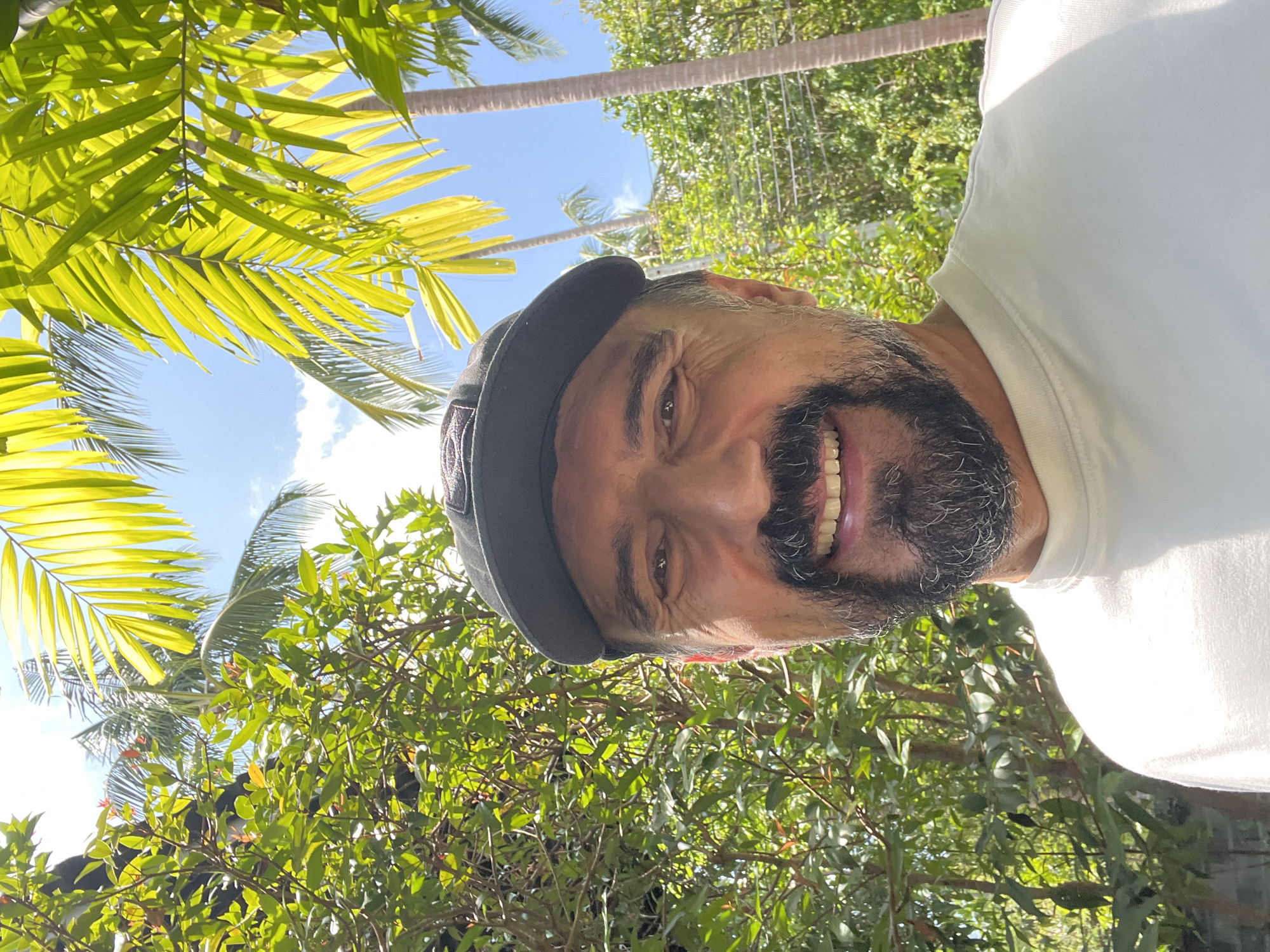 A man with a gray and white beard and gray cap is smiling and looking at the camera. He is wearing a white shirt. The background consists of bright green tropical foliage, including palm leaves, and a blue sky. This is in the city of บ้านบางรักษ์, Thailand.