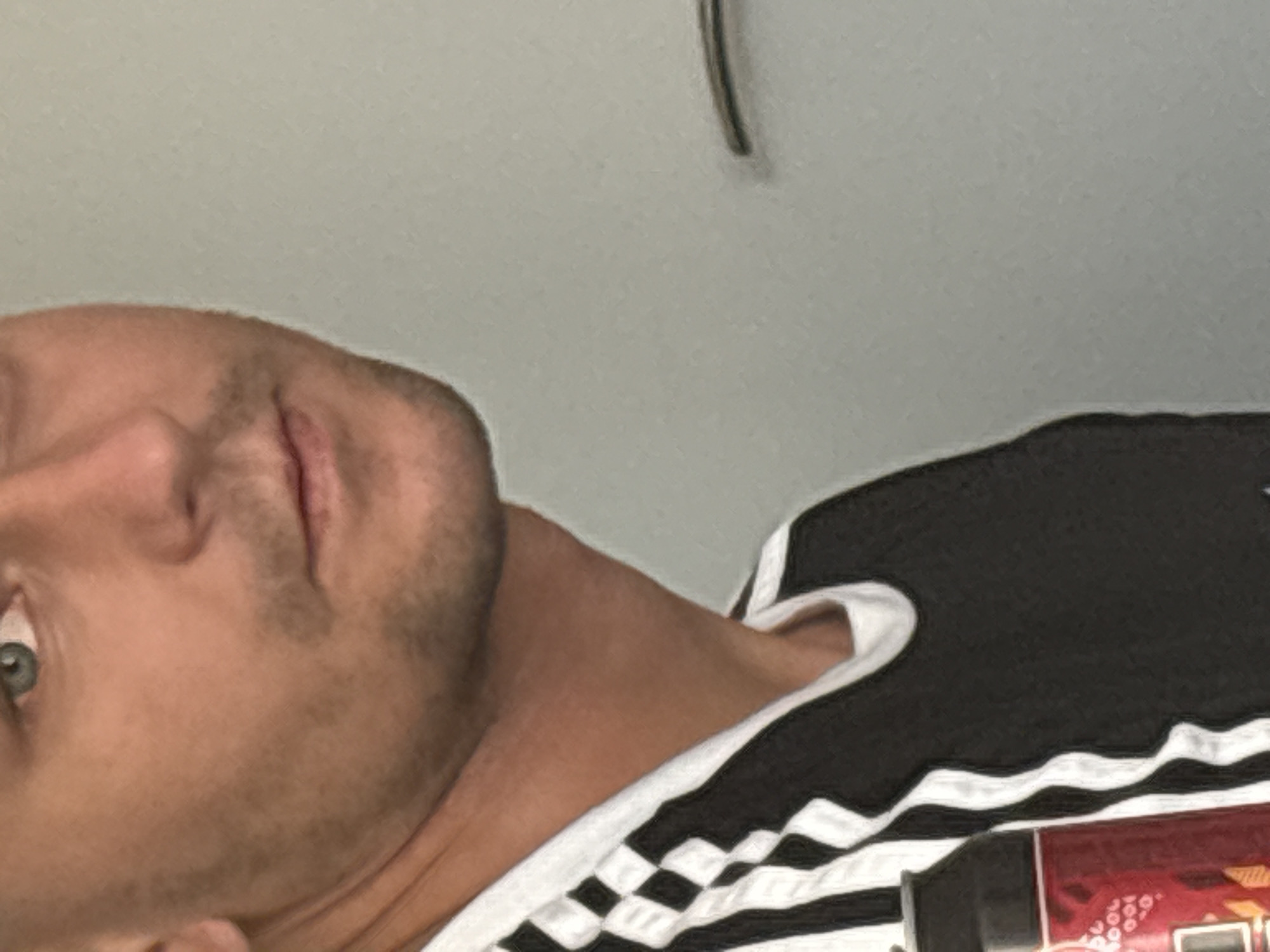 This is a close-up, vertically oriented photograph of a person's face and upper body. The person appears to be a light-skinned adult, with visible stubble on their chin and jawline. Their left eye, which is a light green or hazel color, is clearly visible. The person's mouth is slightly open, revealing their teeth and part of their tongue. They are wearing a black t-shirt with white stripes along the sleeves and neckline. The shirt has a distinctive checkered pattern on the white stripes. The background is a plain, light gray wall. The lighting appears to be indoor, with no clear indication of time of day or weather conditions. There is a faint shadow of an object hanging from the ceiling in the upper right portion of the image. The overall mood of the image is neutral, with no strong emotional cues. No visible text is present in the image.