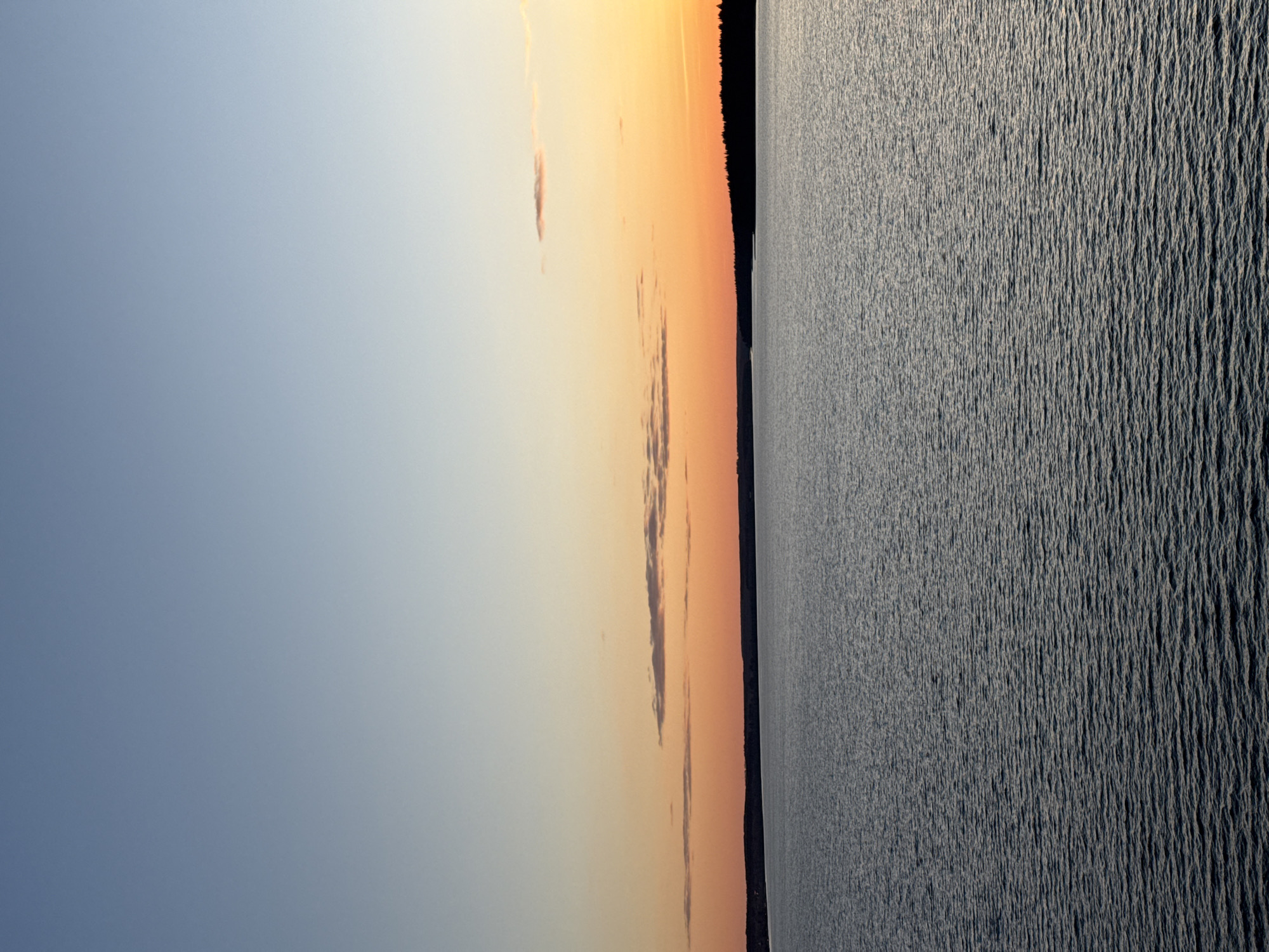 The image depicts a waterfront scene, likely near Votkinsk, Russia, at dusk or dawn. To the left, the sky transitions from blue to orange, with a few elongated clouds scattered across it. A dark, dense treeline separates the sky from the water's edge. The water itself occupies the right portion of the image and displays a textured surface with ripples or small waves. The lighting suggests that the sun is either setting or rising behind the treeline, casting a dark silhouette against the colorful sky.