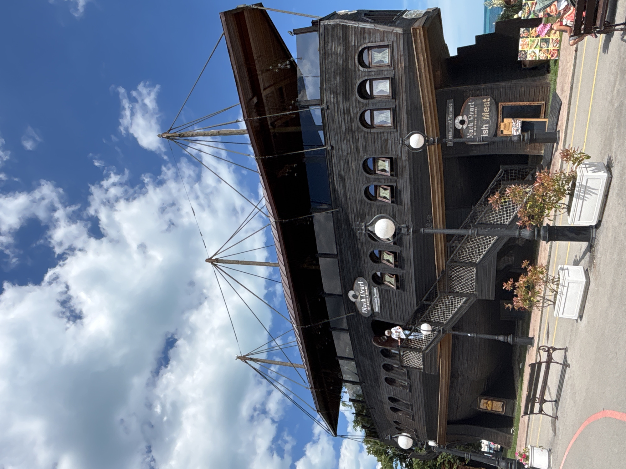 The image depicts a restaurant named "Black Pearl" designed to resemble a pirate ship, located in Varna, Bulgaria. The structure is dark wood with multiple decks and simulated masts and rigging. An elevated entrance features a staircase with rope railings, and a person is seen ascending it. Potted plants and street lamps are positioned along the walkway in front of the restaurant. In the background, a bright blue sky with scattered white clouds is visible, and beyond the immediate foreground, the sea can be seen. The overall scene suggests a sunny day at a seaside resort.